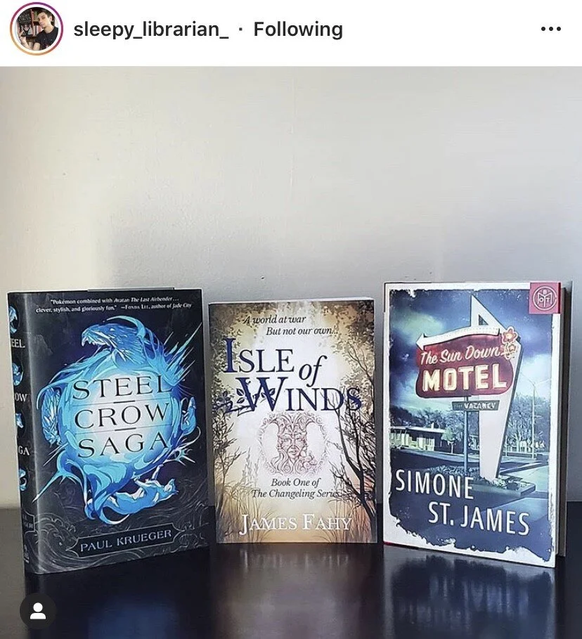 Three books standing upright on a dark surface against a plain wall. The first book is titled 'Steel Crow Saga' by Paul Krueger, the second is 'Isle of Winds' by James Fahy, and the third is 'The Sun Down Motel' by Simone St. James.