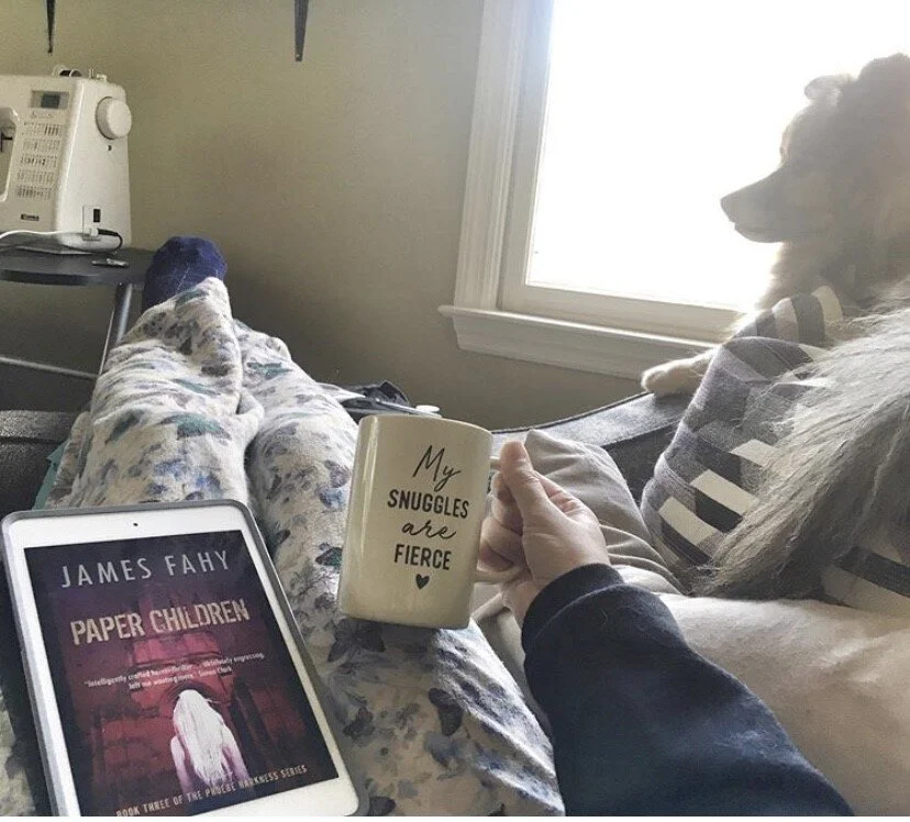 A person sitting on a couch holding a mug that reads 'My snuggles are fierce' while reading a book titled 'Paper Children' by James Fahy. There is a dog sitting beside the person and a tablet on their lap displaying the book cover. The person is wear