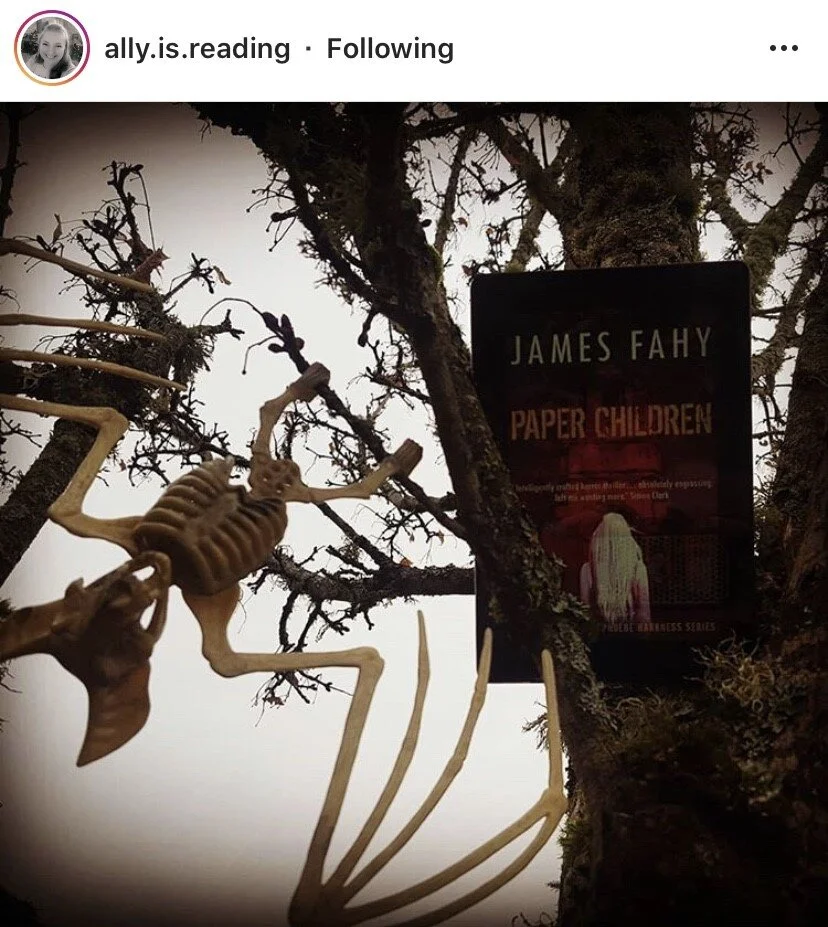 A piece of a skeleton model or toy hanging from a leafless tree branch with a book titled "Paper Children" by James Fahy attached to the tree, with a woman with long blonde hair on the cover.