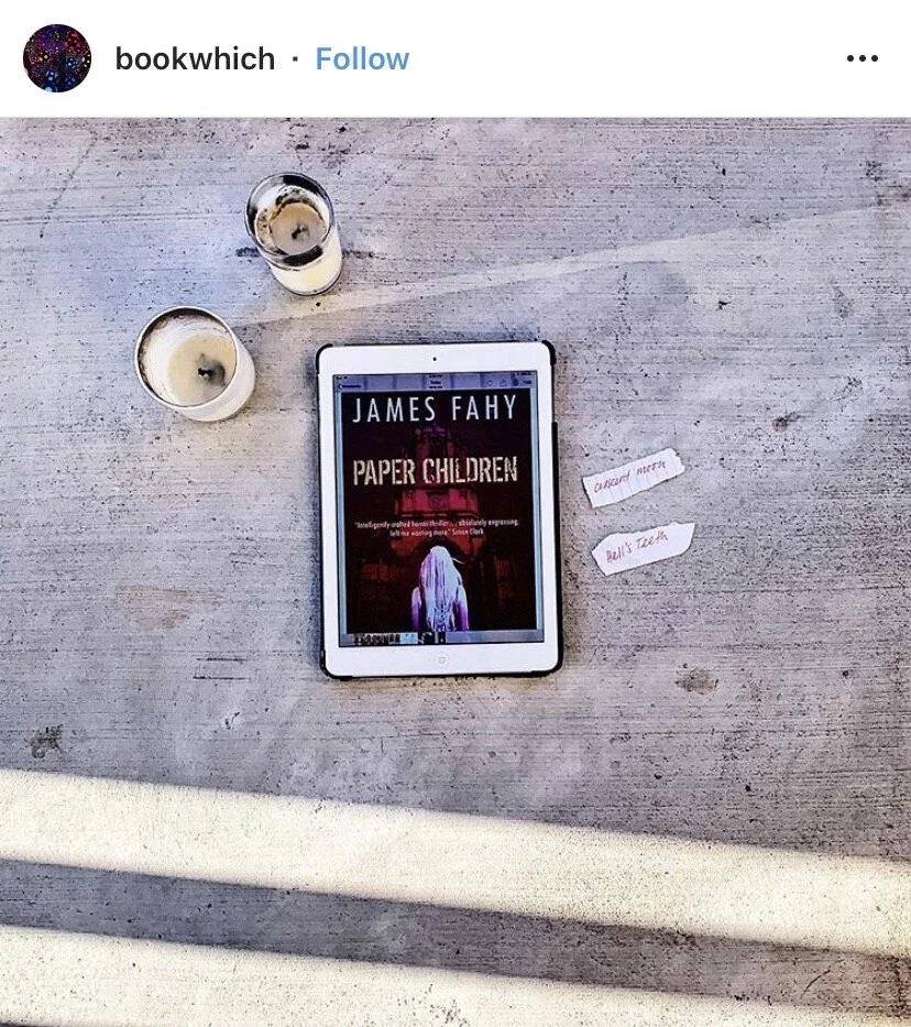 An e-reader displaying the book cover of "Paper Children" by James Fahy, with two candles and two small torn paper pieces next to it reading "on second thought" and "Dell's Team" on a wooden surface.