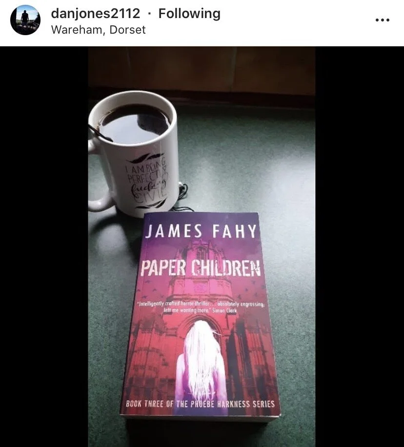 A white mug filled with coffee and a purple book titled "Paper Children" by James Fahy on a green surface, with a background of a wooden surface.