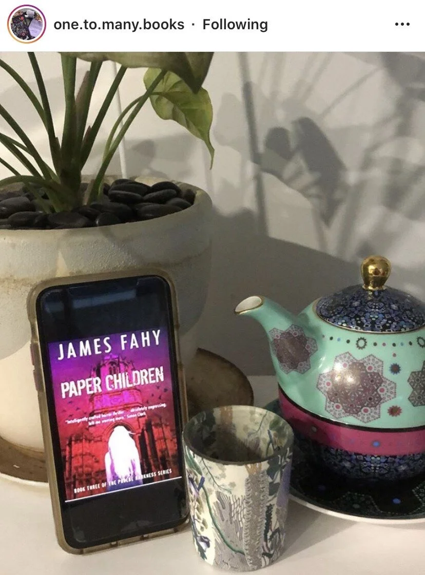 A potted plant, a teapot with colorful geometric patterns, a mug with abstract designs, and a smartphone displaying the cover of the book "Paper Children" by James Fahy.