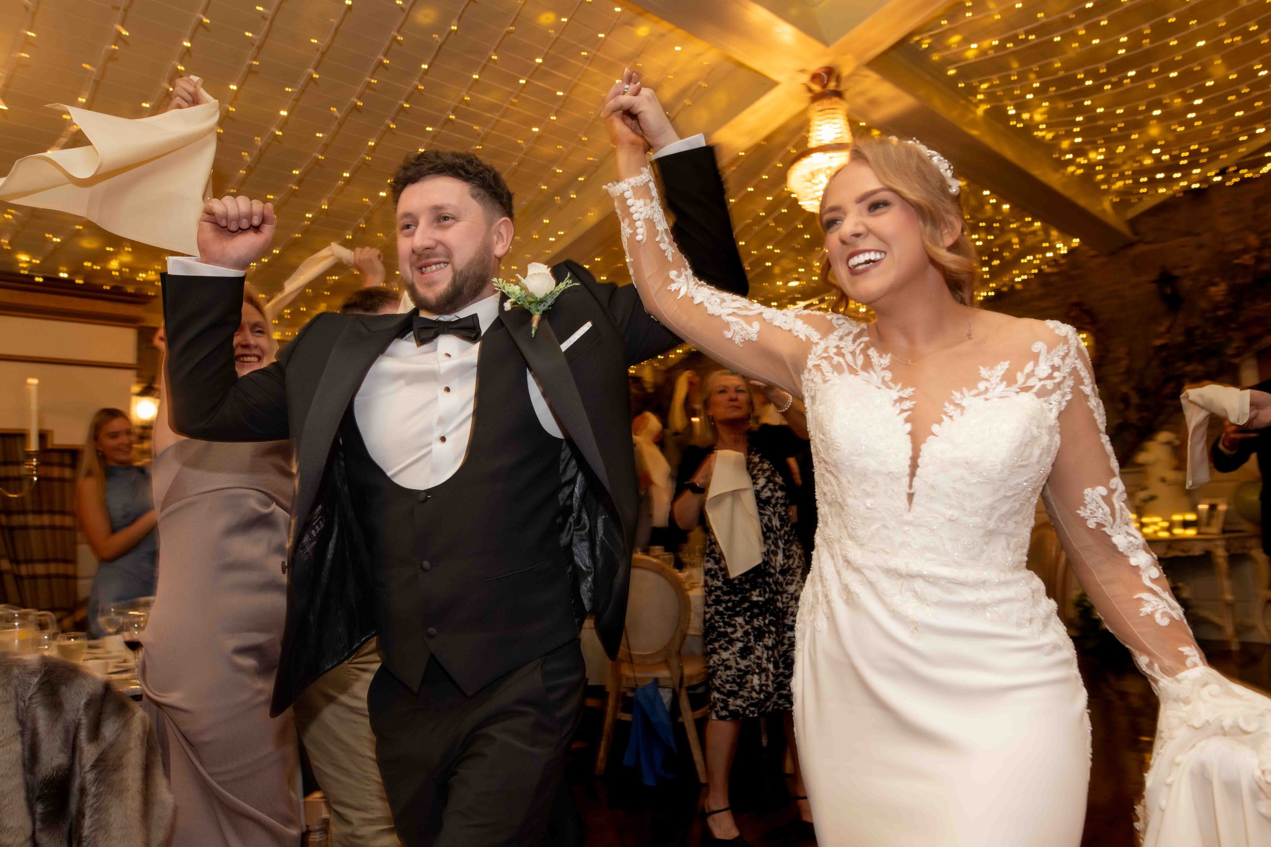 Bride and groom celebrating at a wedding reception with guests cheering under twinkling lights.