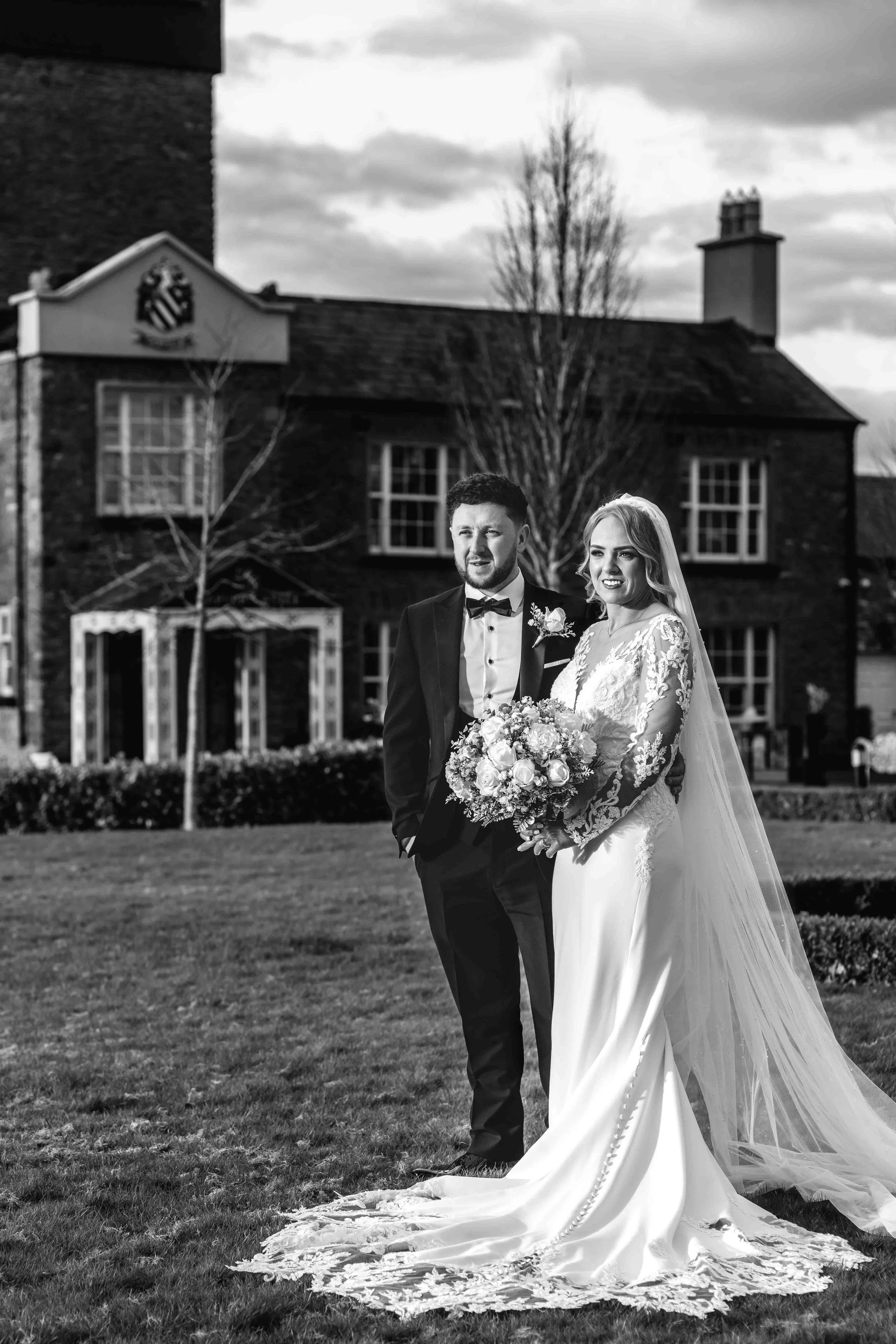 Bride and groom posing outdoors in wedding attire, with a building in the background.