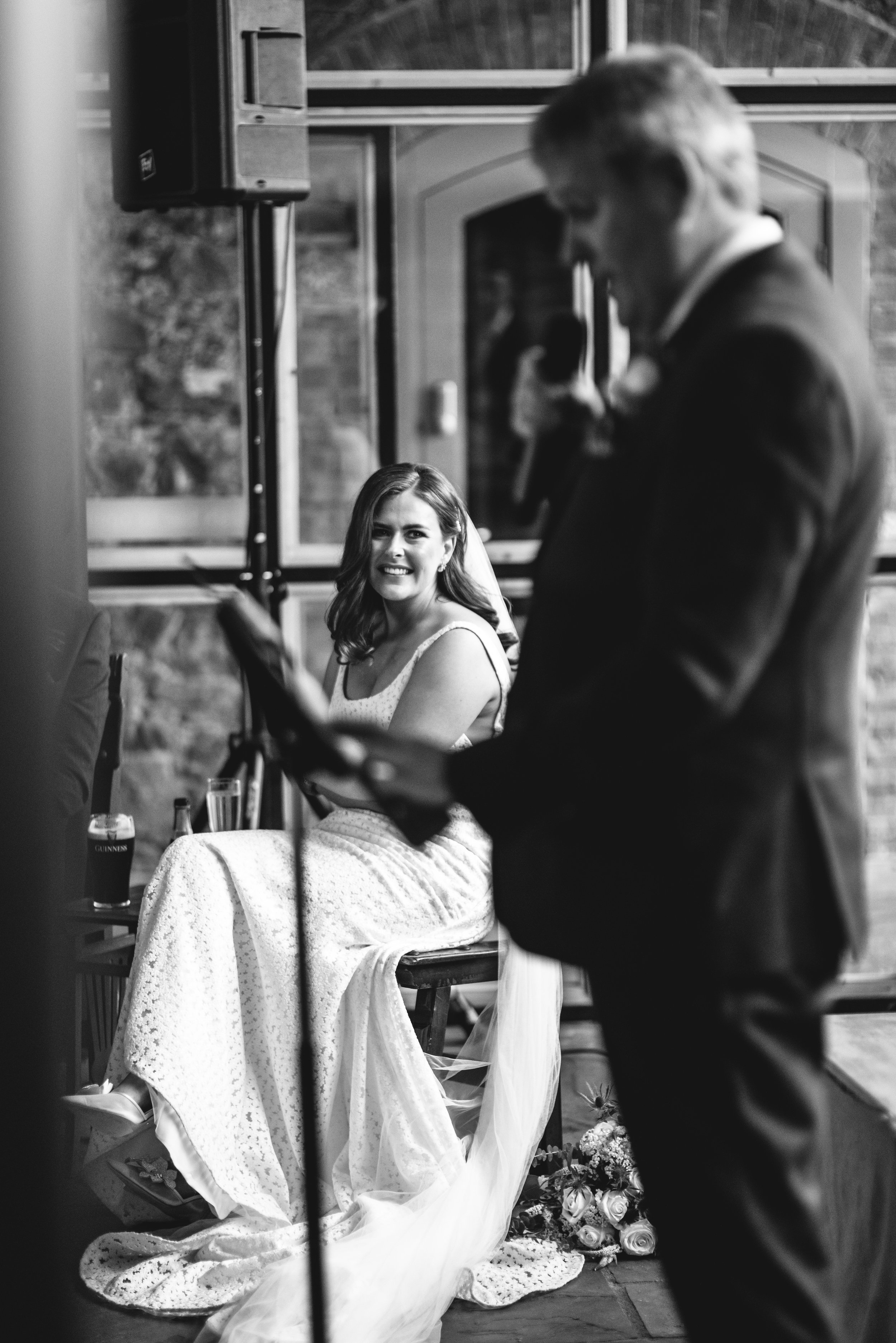 Bride sitting in a wedding ceremony, listening to a speech by a man holding a microphone, with flowers on the floor beside her.