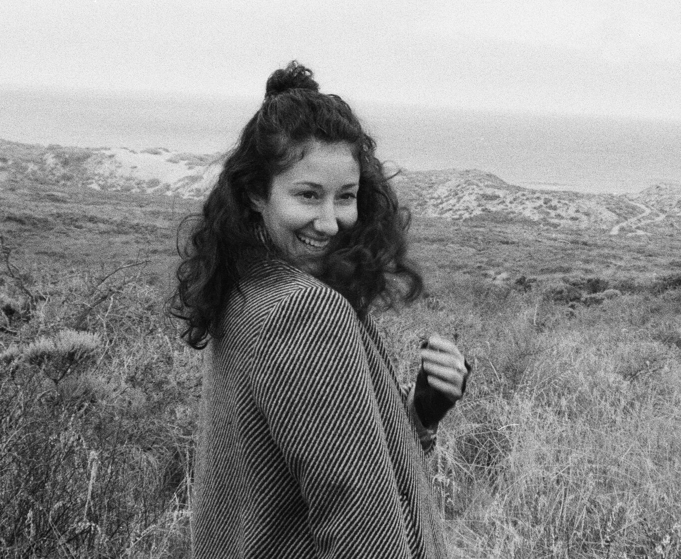 A smiling woman with brown curly hair outdoors in a sand dune landscape, wearing a striped jacket.