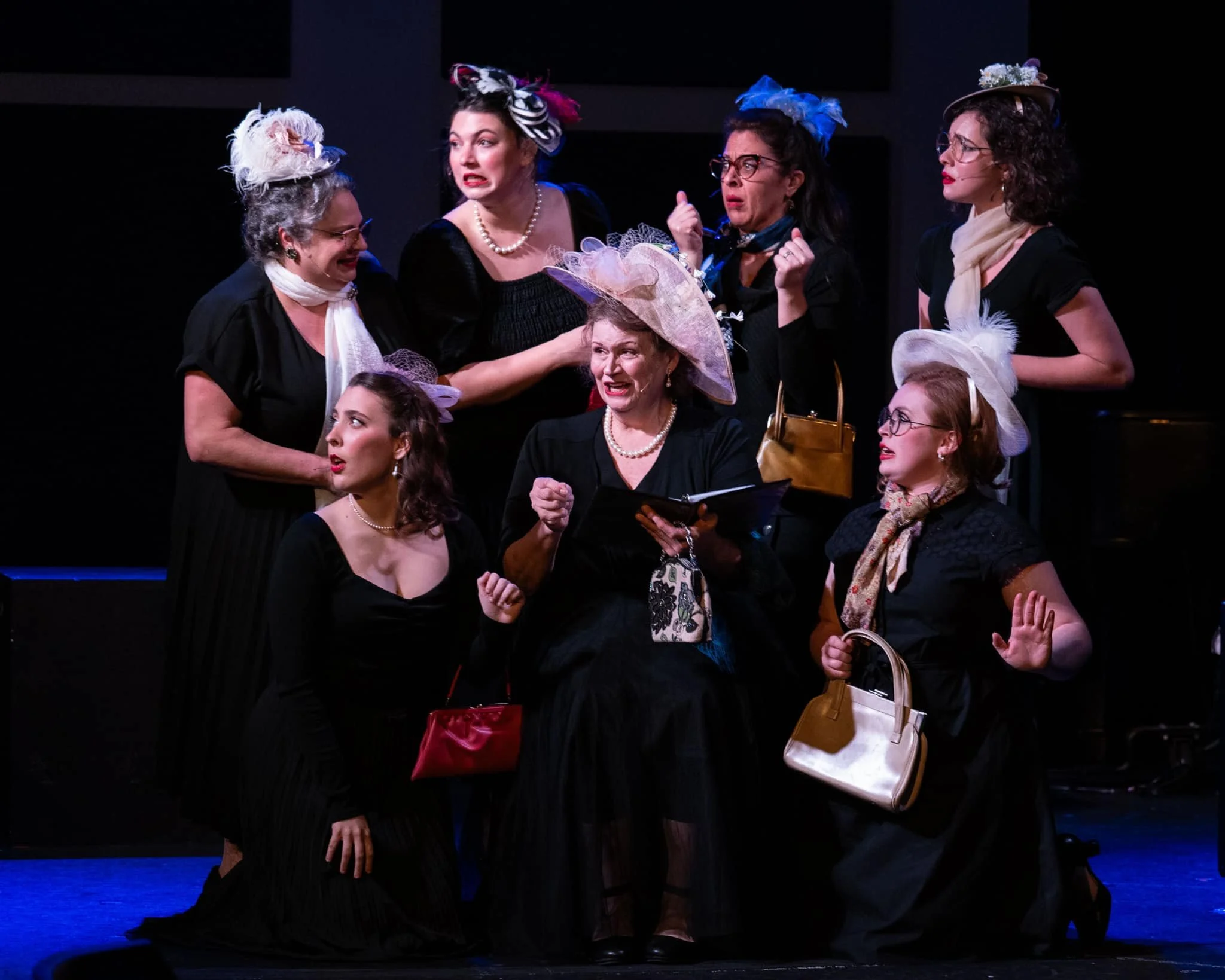 A theatrical performance with seven women dressed in vintage-style clothing, including black dresses and hats decorated with feathers, flowers, and ribbons, gathered on stage against a black background.