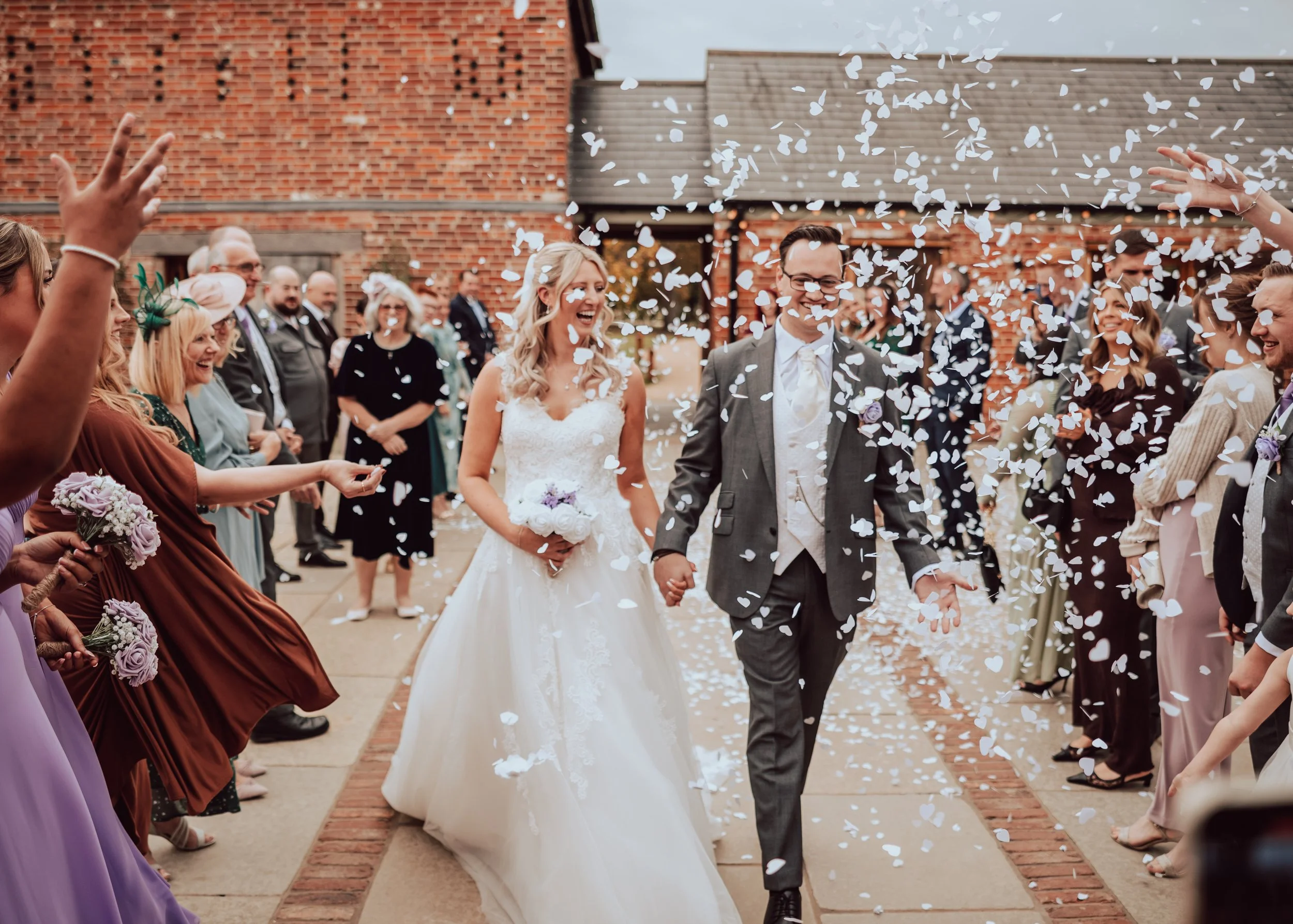 A bride and groom walking hand-in-hand during their wedding celebration, surrounded by guests throwing white confetti outside near a brick building.