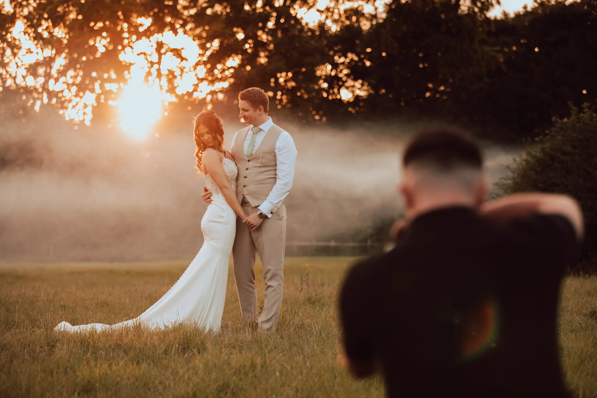 A bride and groom standing close together in a field at sunset, with a photographer capturing their wedding moments. The bride is in a white wedding gown, and the groom is in a light-colored suit. The background features trees illuminated by the setting sun.