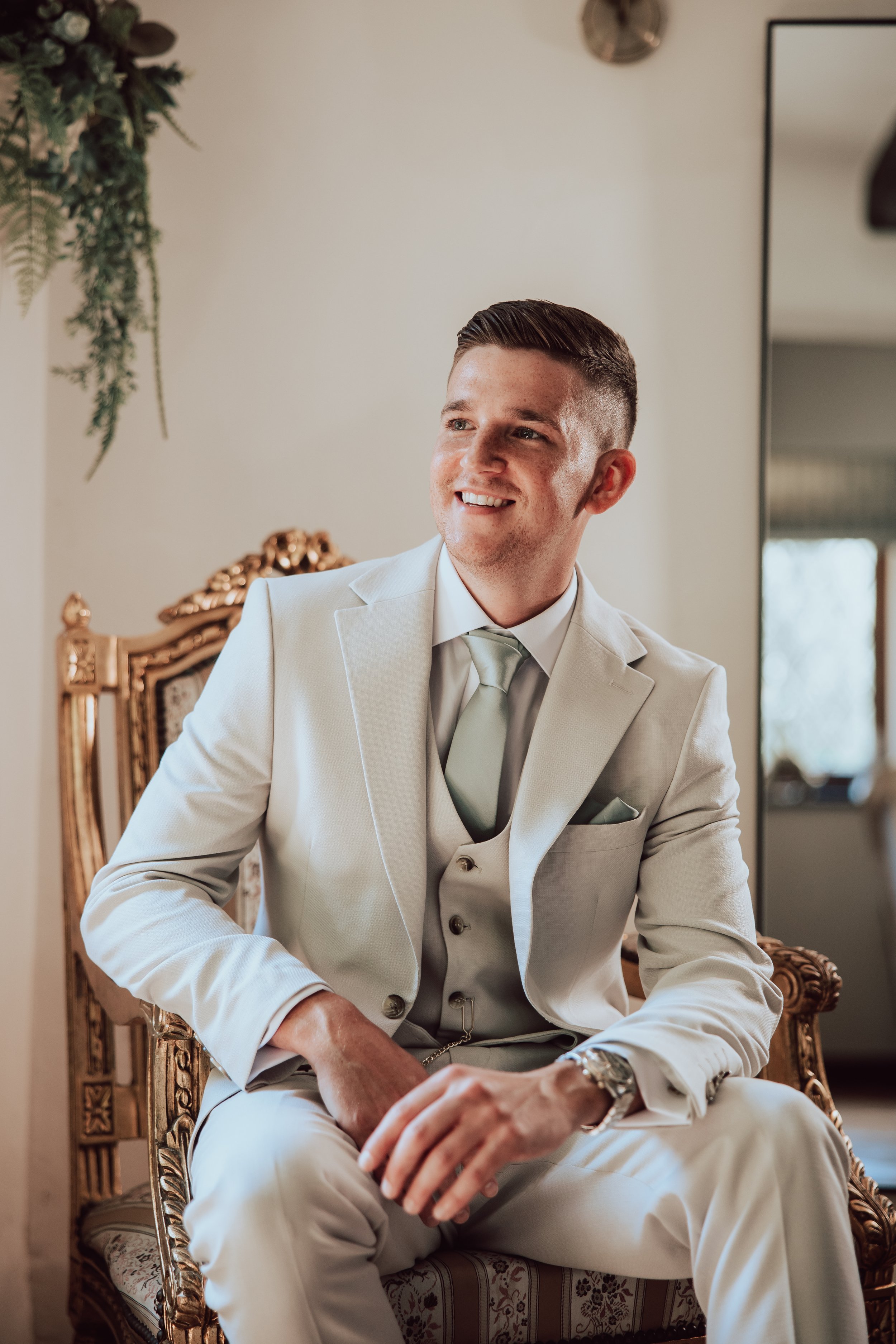 A man dressed in a light-colored suit with a tie, sitting on an ornate wooden chair with a patterned cushion, smiling and looking to his left in a well-lit room.