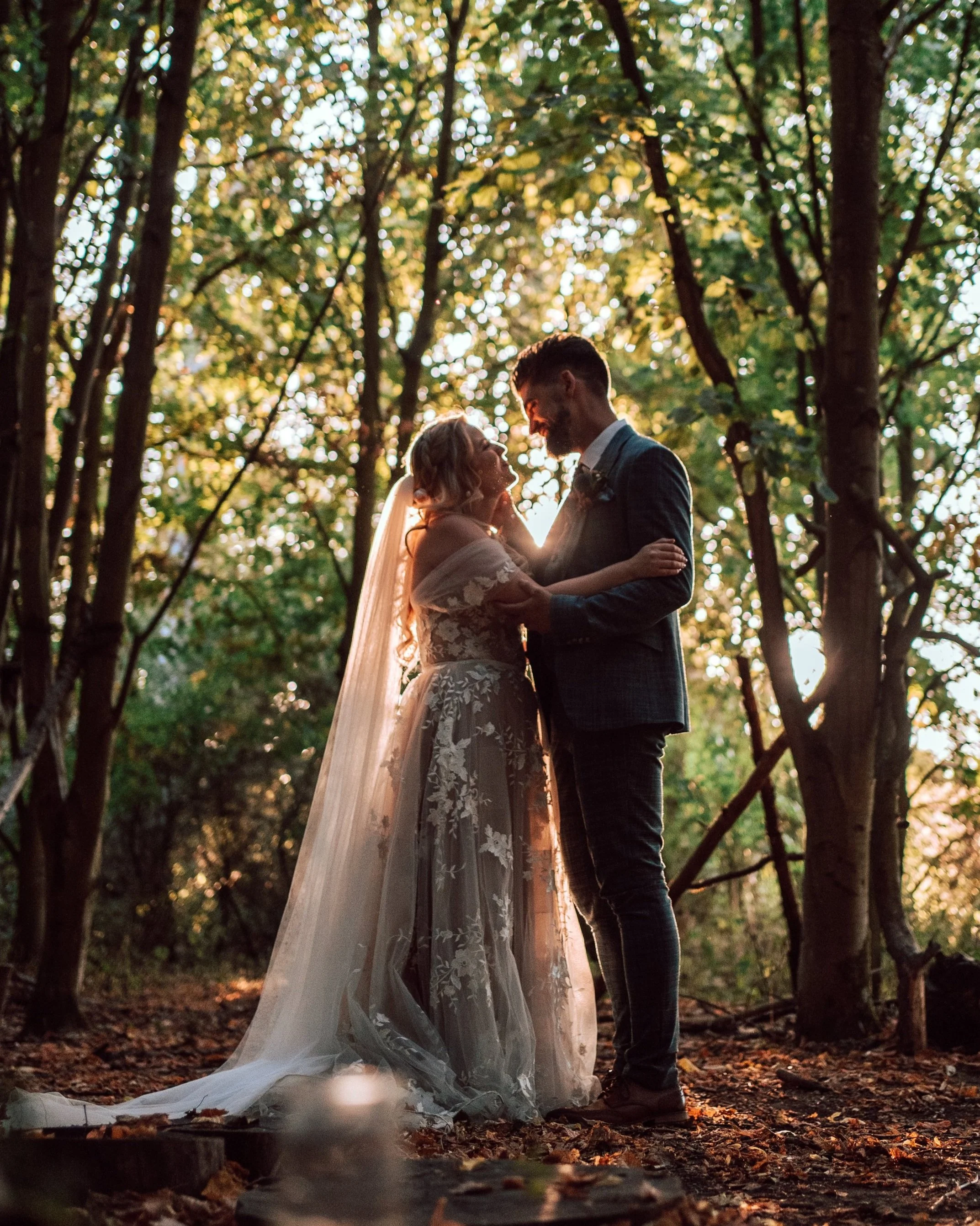 A bride and groom smiling and looking at each other while holding each other close in a wooded area during sunset, with dappled sunlight coming through the trees.