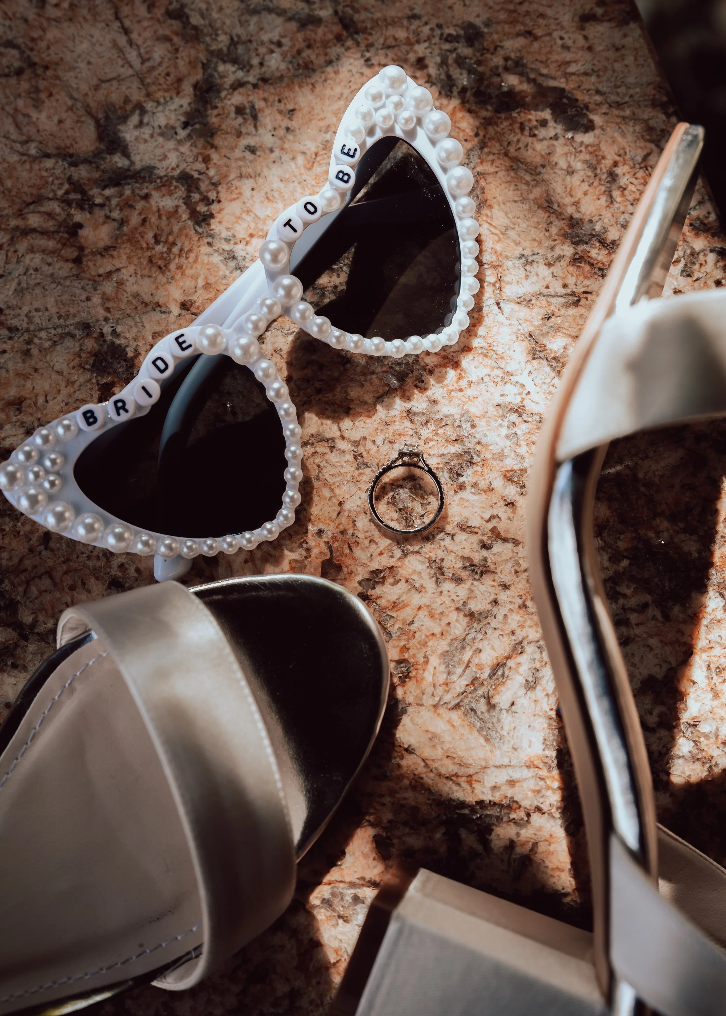 Pearl-beaded heart-shaped sunglasses with the words 'BRIDE TO BE' on the frame, sitting on a granite countertop near a pair of silver high heels.