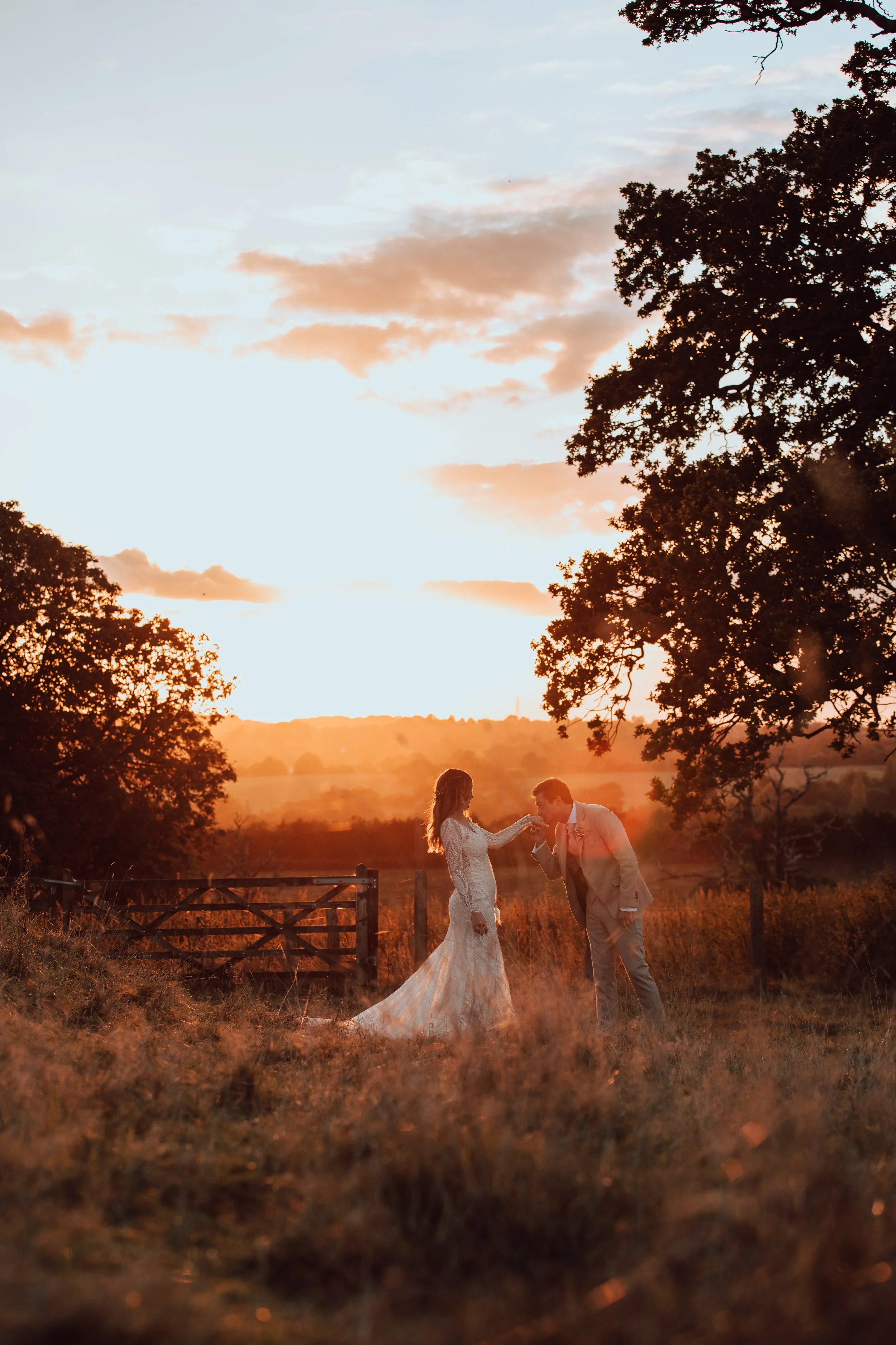 A couple in wedding attire exchanging vows outdoors at sunset, with trees and a rural landscape in the background.