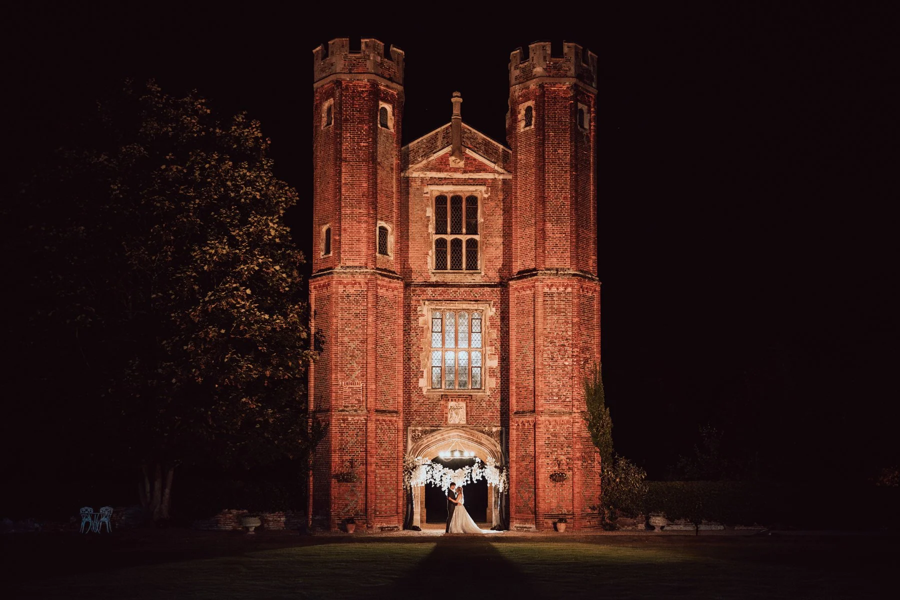 Nighttime wedding scene with a bride and groom standing beneath a lit church archway, in front of a historic red brick church with two tall towers, surrounded by trees.