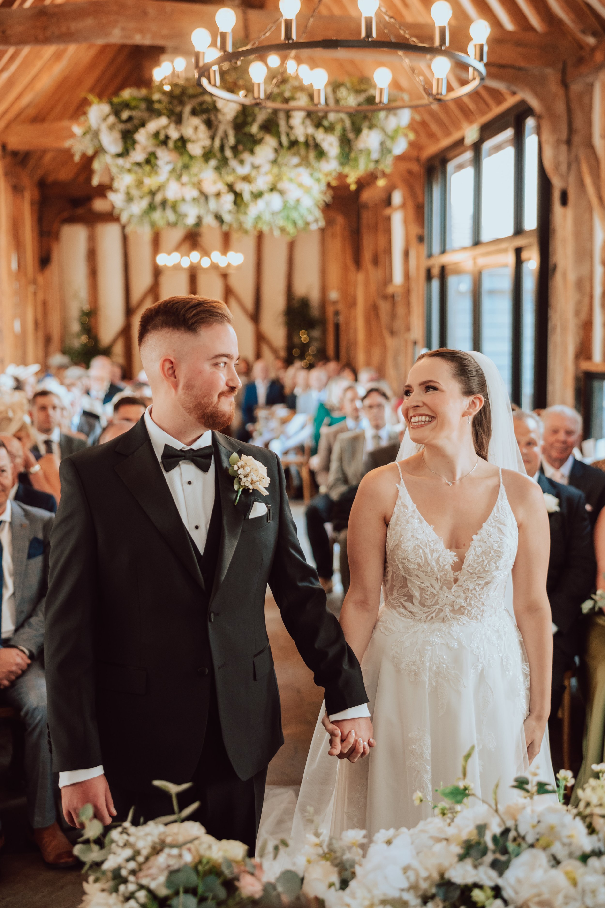 A bride and groom holding hands during their wedding ceremony inside a rustic wooden venue, surrounded by wedding guests and floral decorations.