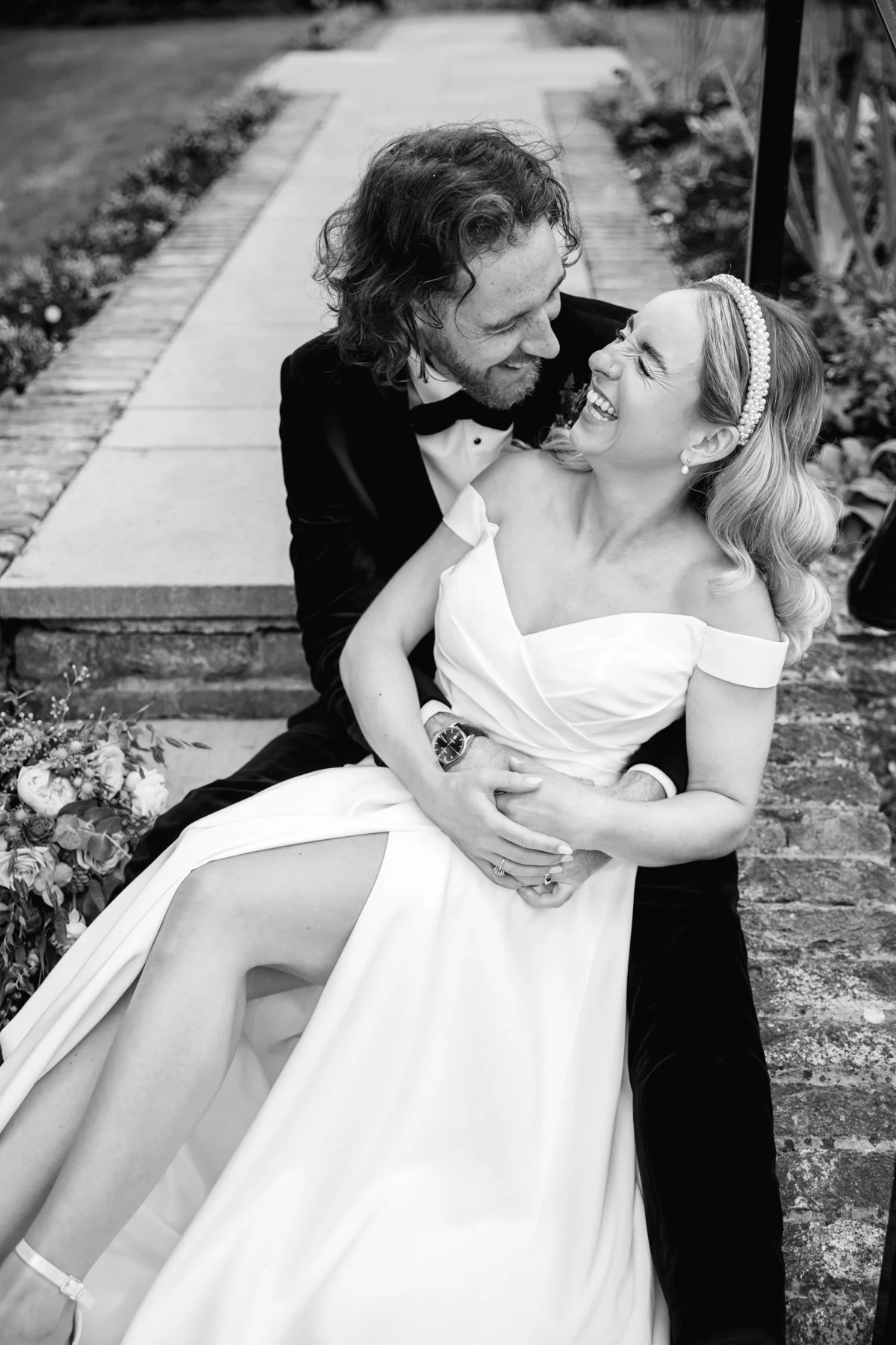 Black and white photo of a joyful couple, with the man in a tuxedo and the woman in a wedding dress, sharing a happy moment outdoors on a sidewalk.