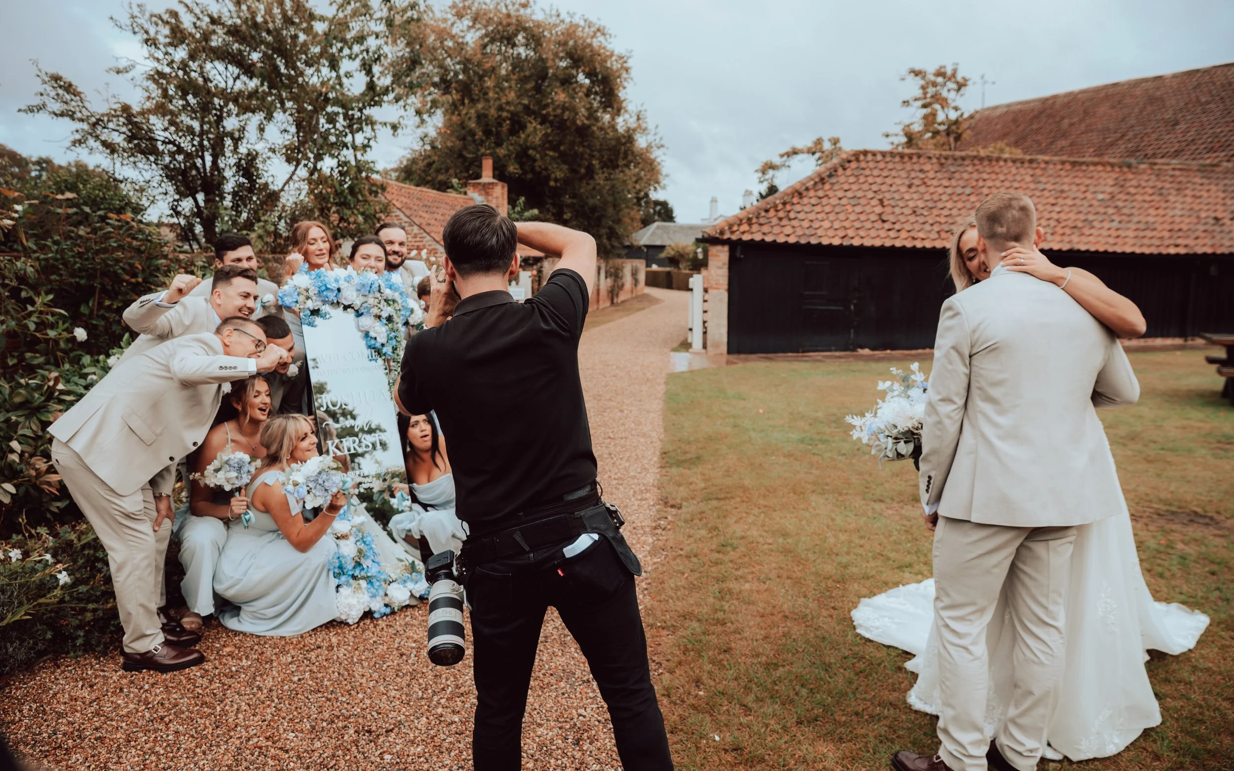 A wedding celebration outdoors with a group of bridesmaids and groomsmen, some holding bouquets, taking photos of the bride and groom embracing in front of a decorated seating area with a sign, across a gravel path, with trees and a rustic building with a tiled roof in the background.