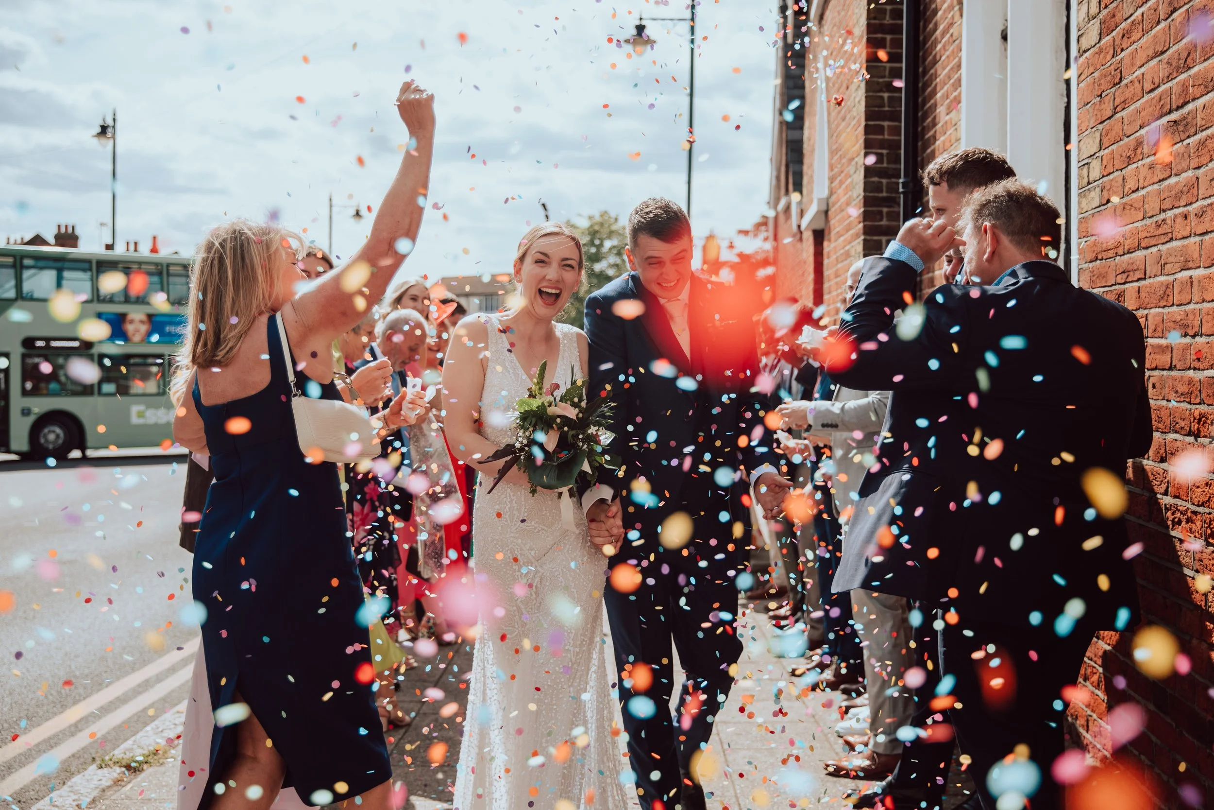 A group of people celebrating a wedding outside on a street, surrounded by colorful confetti. The bride and groom are holding hands and smiling, with the bride holding a bouquet. Guests are cheering and throwing confetti, and the background shows a brick building and a bus.