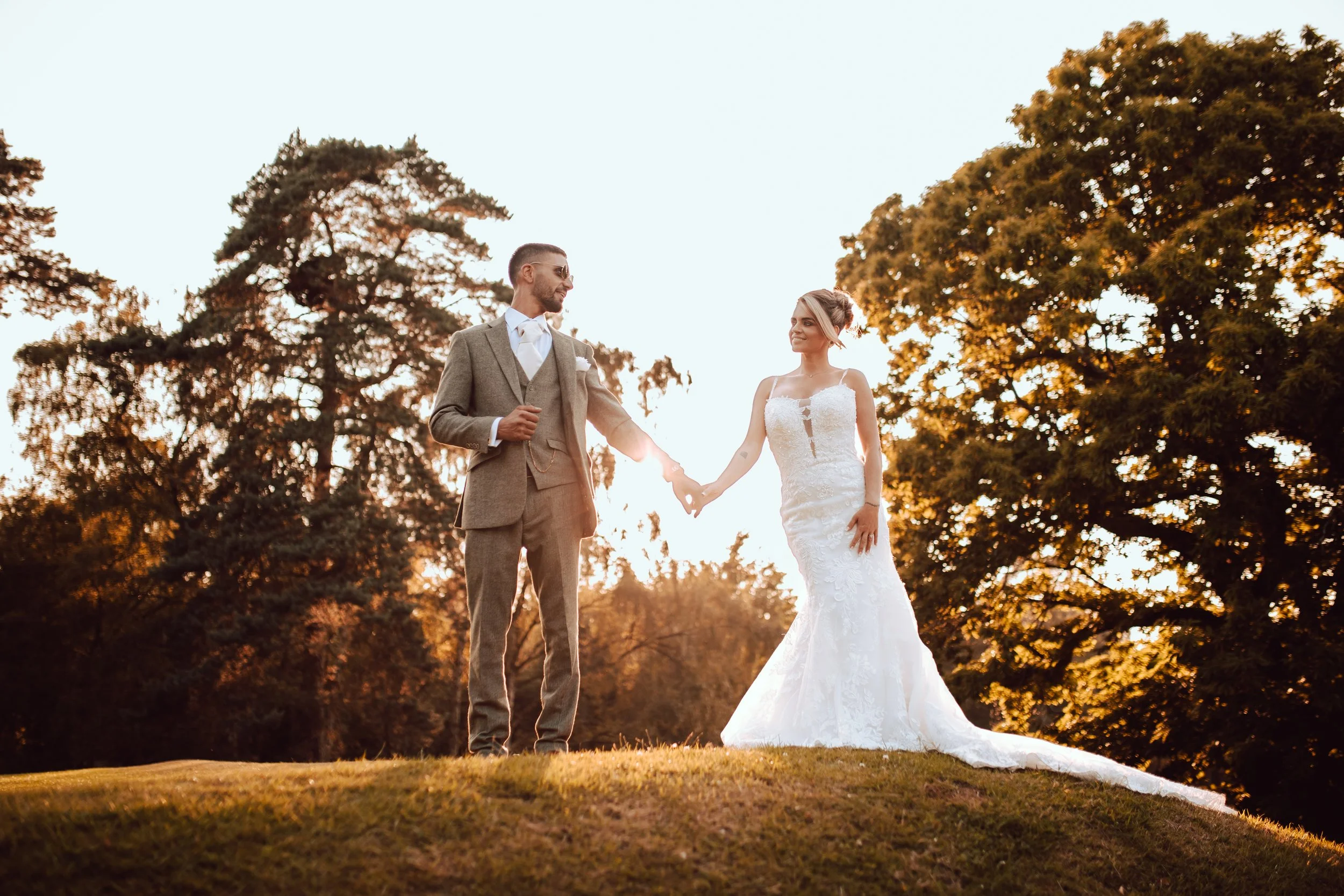 A wedding couple standing outdoors during sunset, holding hands and smiling. The groom is wearing a gray suit and the bride is in a white lace wedding dress.
