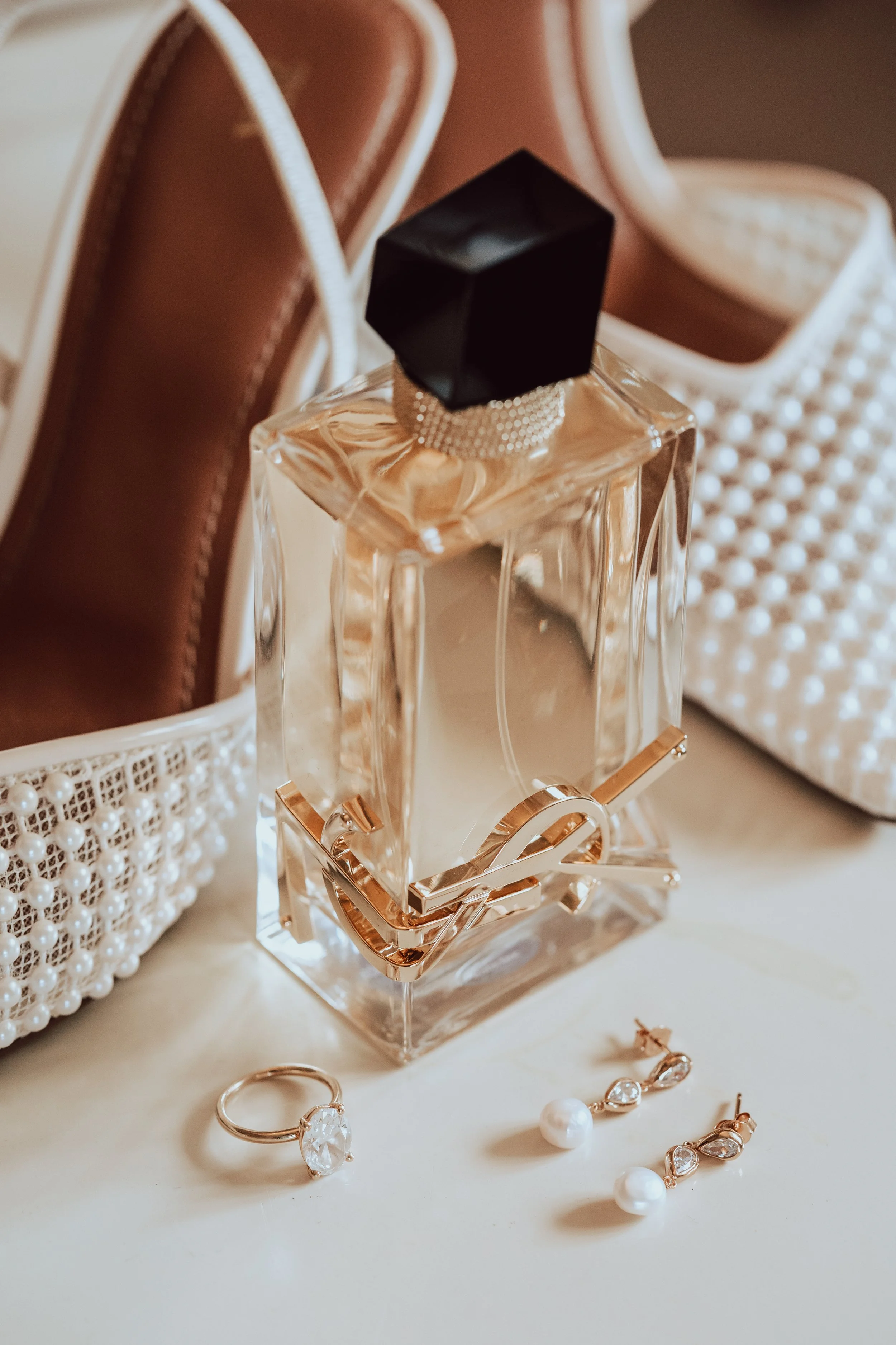 A perfume bottle with a black cap and rose gold accents on a white surface, surrounded by pearl jewelry and decorative boxes.