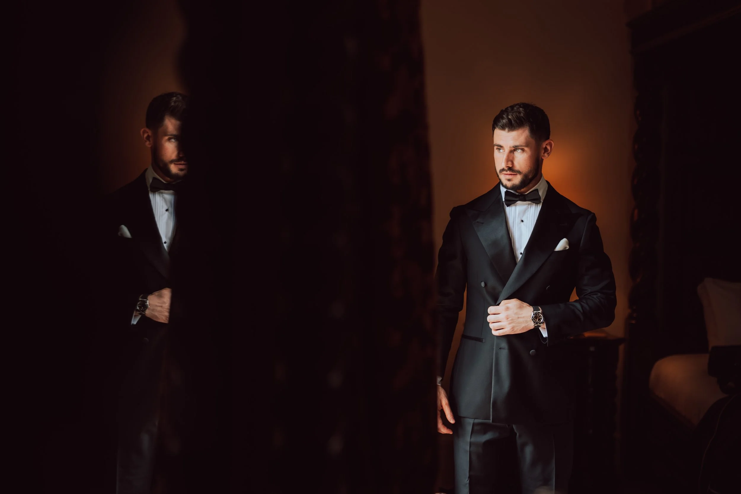 A man in a black tuxedo with a bowtie and watch, looking at his reflection in a mirror in a dimly lit room.