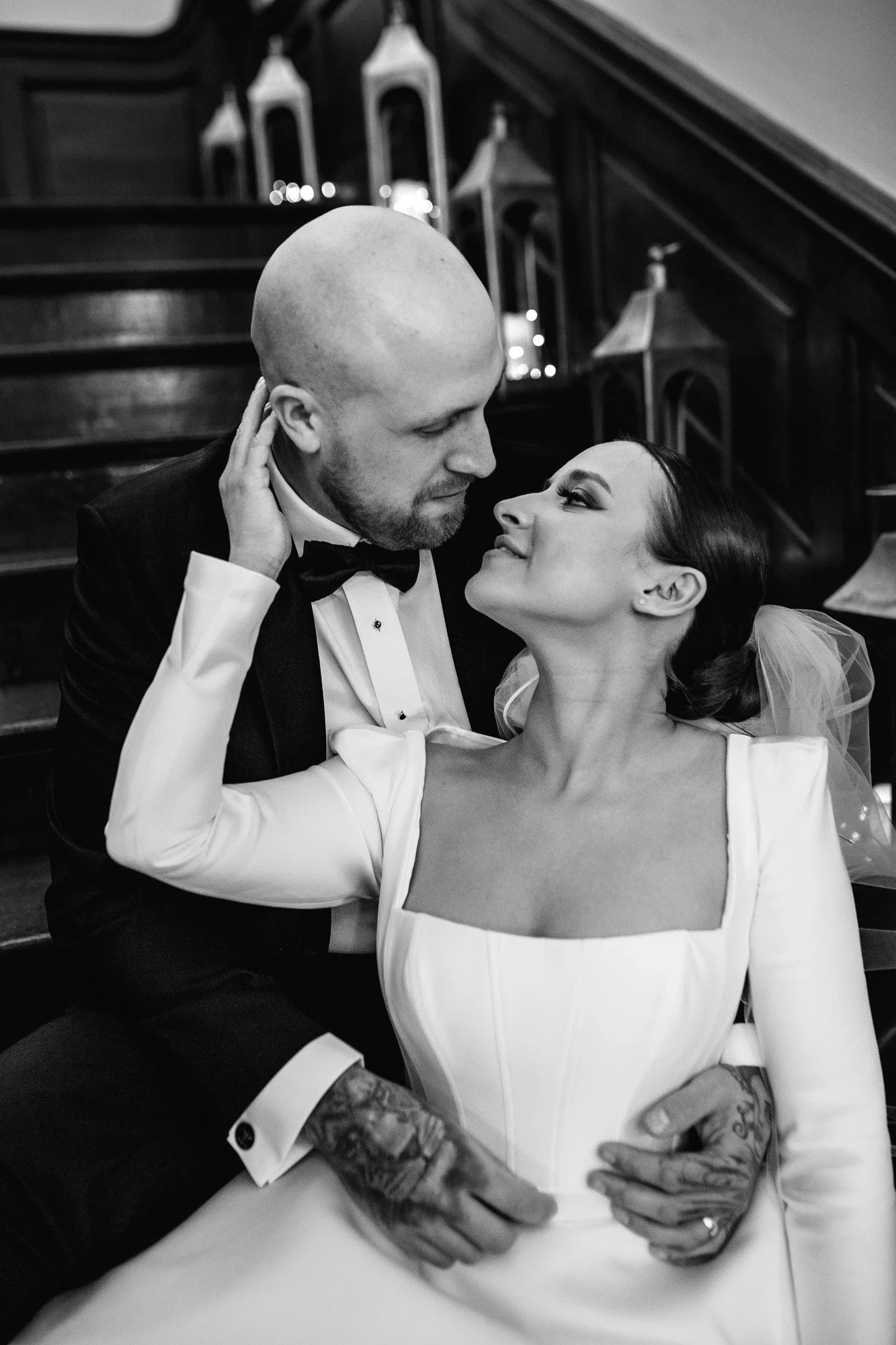 A black and white photo of a couple at their wedding, sitting closely together and looking into each other's eyes. The bride is wearing a white gown with a square neckline and tulle detail, and the groom is in a tuxedo with a bowtie. They are holding hands, displaying tattoos on their wrists and hands, with a wooden staircase and small candles in the background.