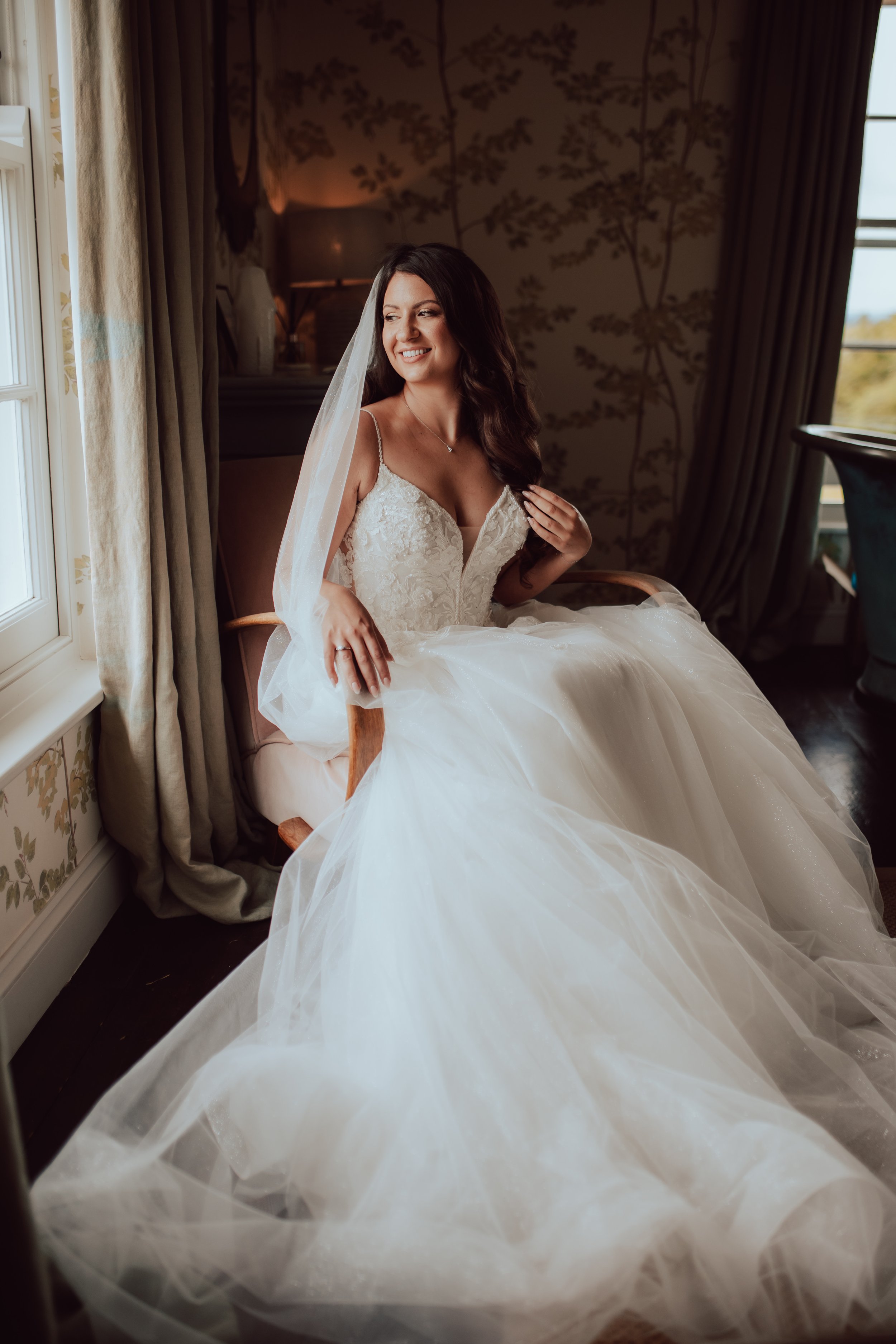 A bride with long dark hair in a white lace wedding dress, sitting in a vintage-style room by a window, with curtains and floral wallpaper, smiling and looking to her left.