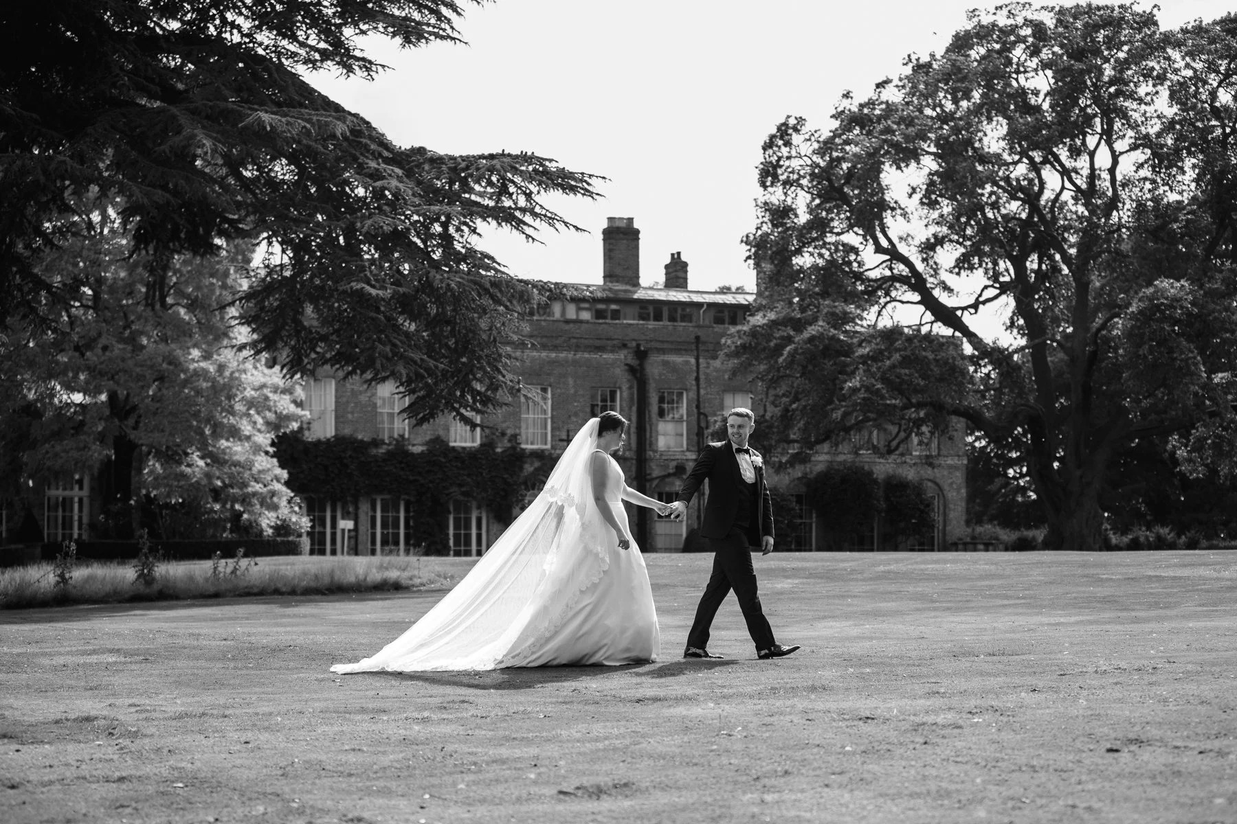 A black and white photo of a bride and groom holding hands, walking on a lawn in front of a large historic building with trees and chimneys.