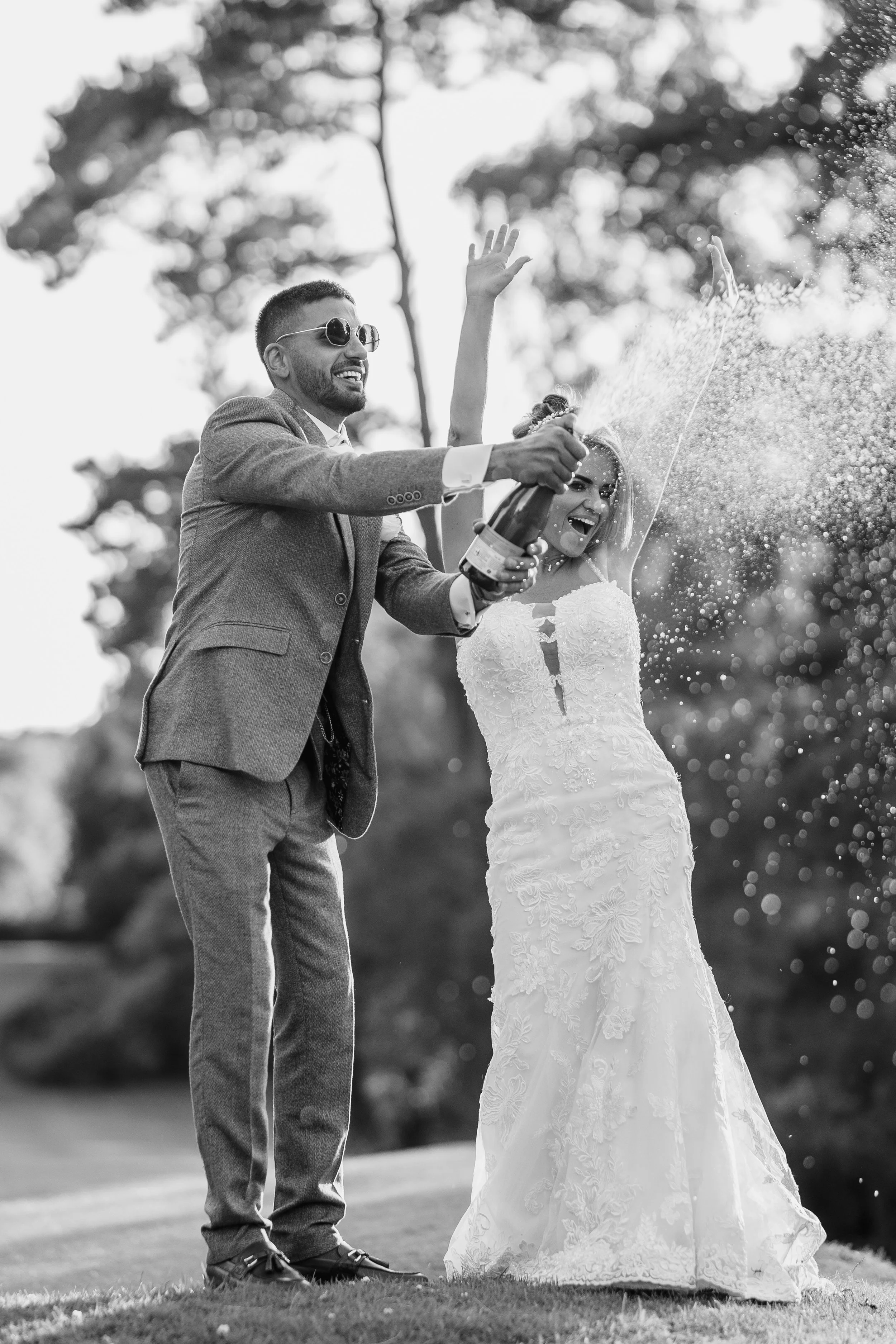 Black and white photo of a wedding celebration featuring a man in a suit and sunglasses holding a champagne bottle, spraying champagne, with a smiling woman in a wedding dress nearby, outdoors with trees in the background.