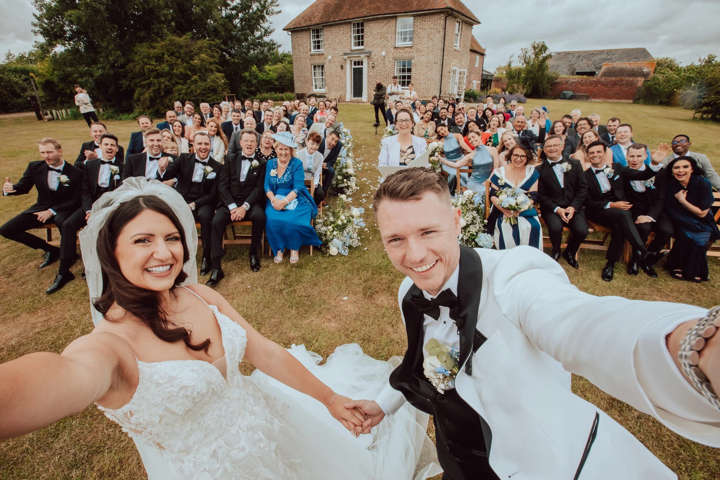 A bride and groom taking a selfie at their outdoor wedding reception, surrounded by large group of wedding guests sitting on chairs on a lawn in front of a brick house.