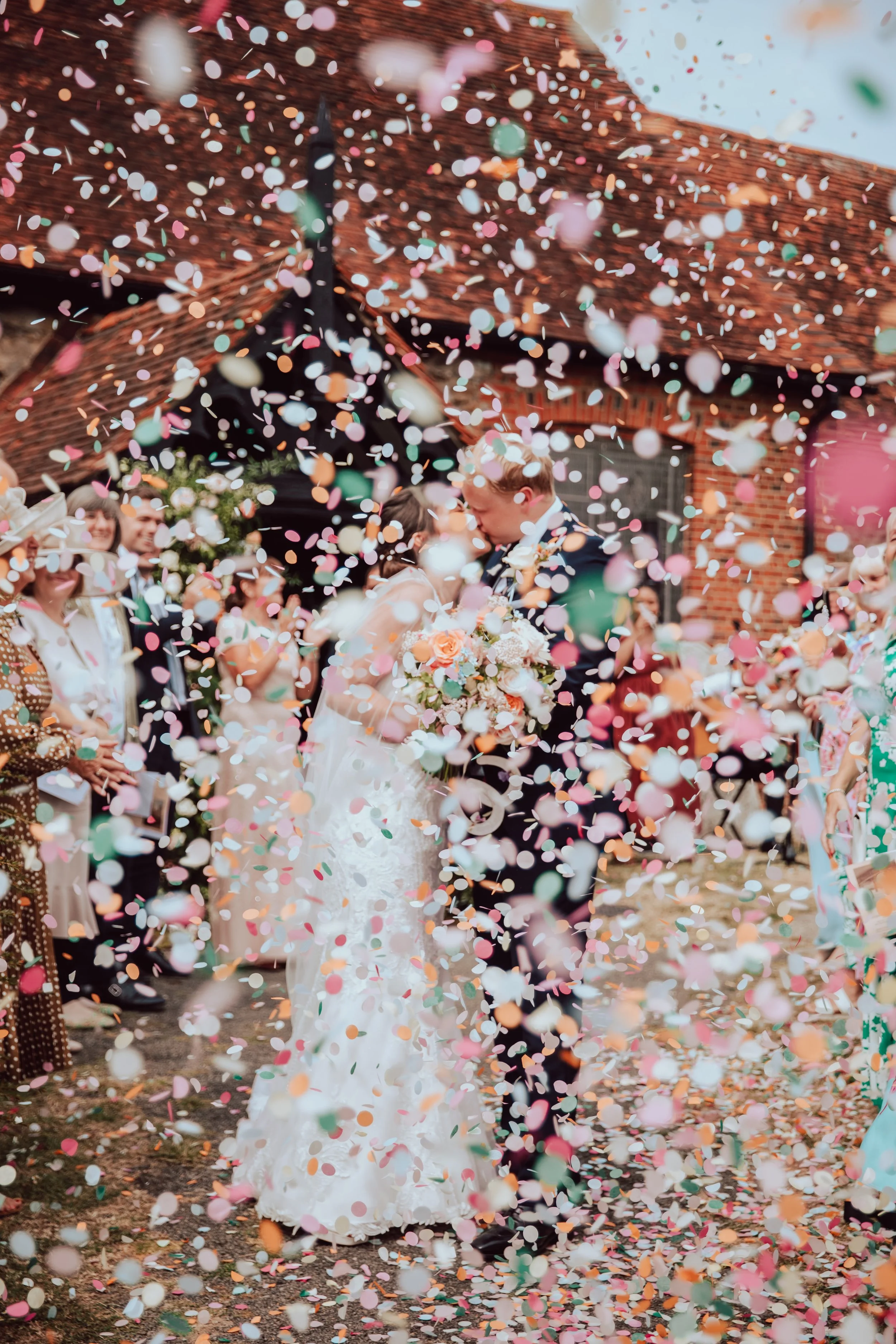 A wedding couple kissing amidst falling pastel-colored confetti with guests smiling in the background outside a brick building.