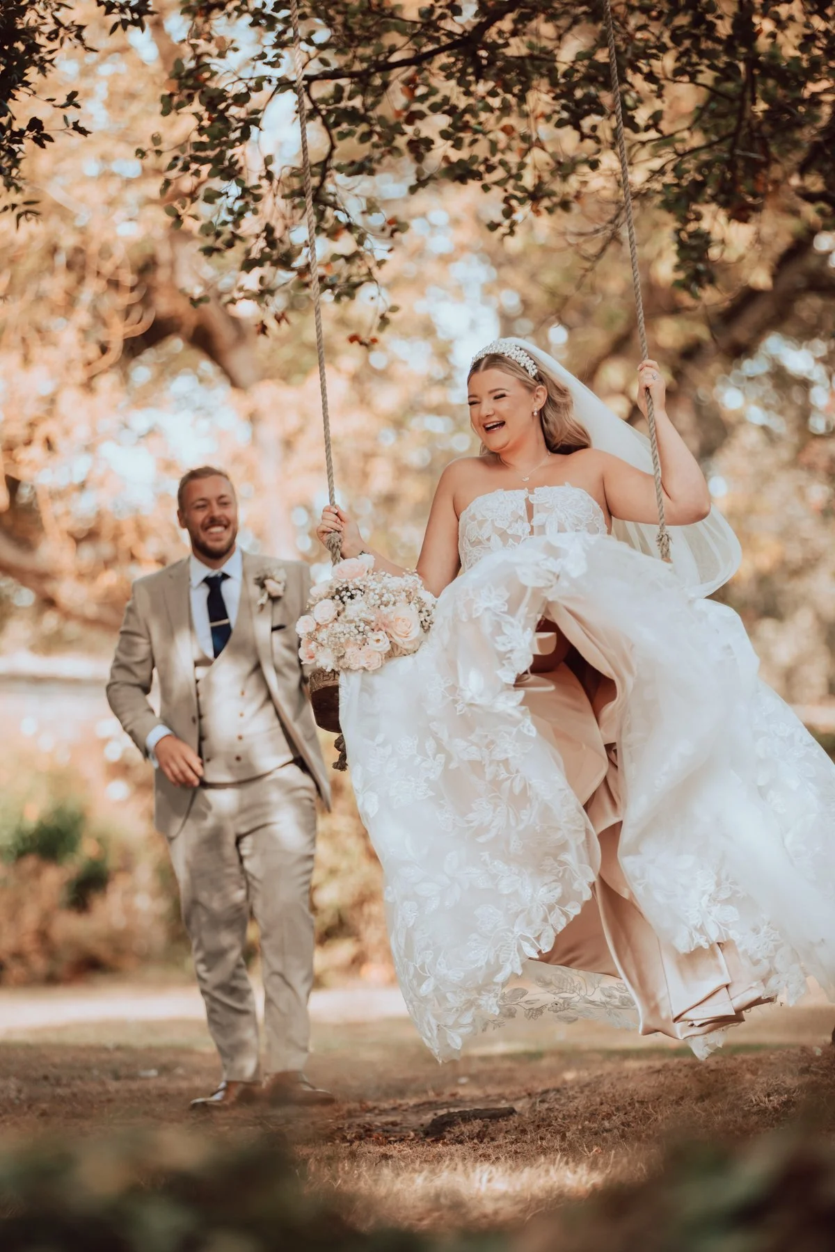 A bride on a swing holding a bouquet of flowers, laughing, with a groom walking behind her, in an outdoor setting with trees and sunlight filtering through the leaves.