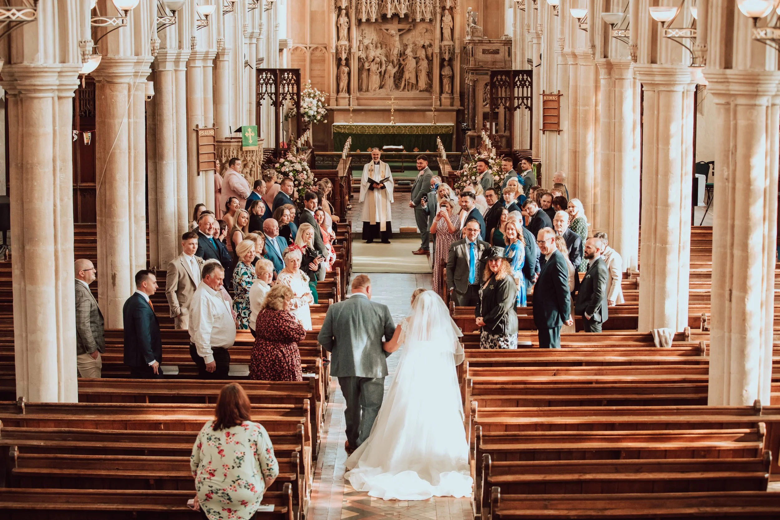 A wedding ceremony taking place inside a large, ornate church with many guests sitting on wooden pews. The bride and groom stand at the front near the altar, with the priest officiating. The church has tall columns and detailed stonework on the walls.