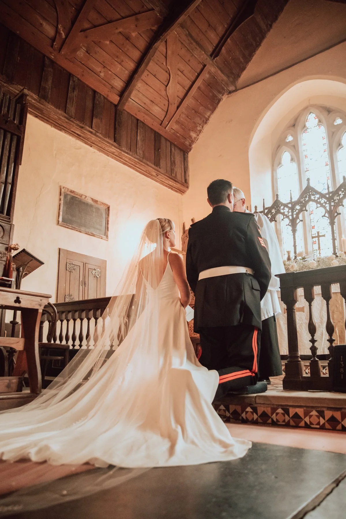A bride in a white wedding dress with a long train and veil stands at the altar in a church, praying in front of an officiant and a man in military uniform. Sunlight streams through stained glass windows in the background.