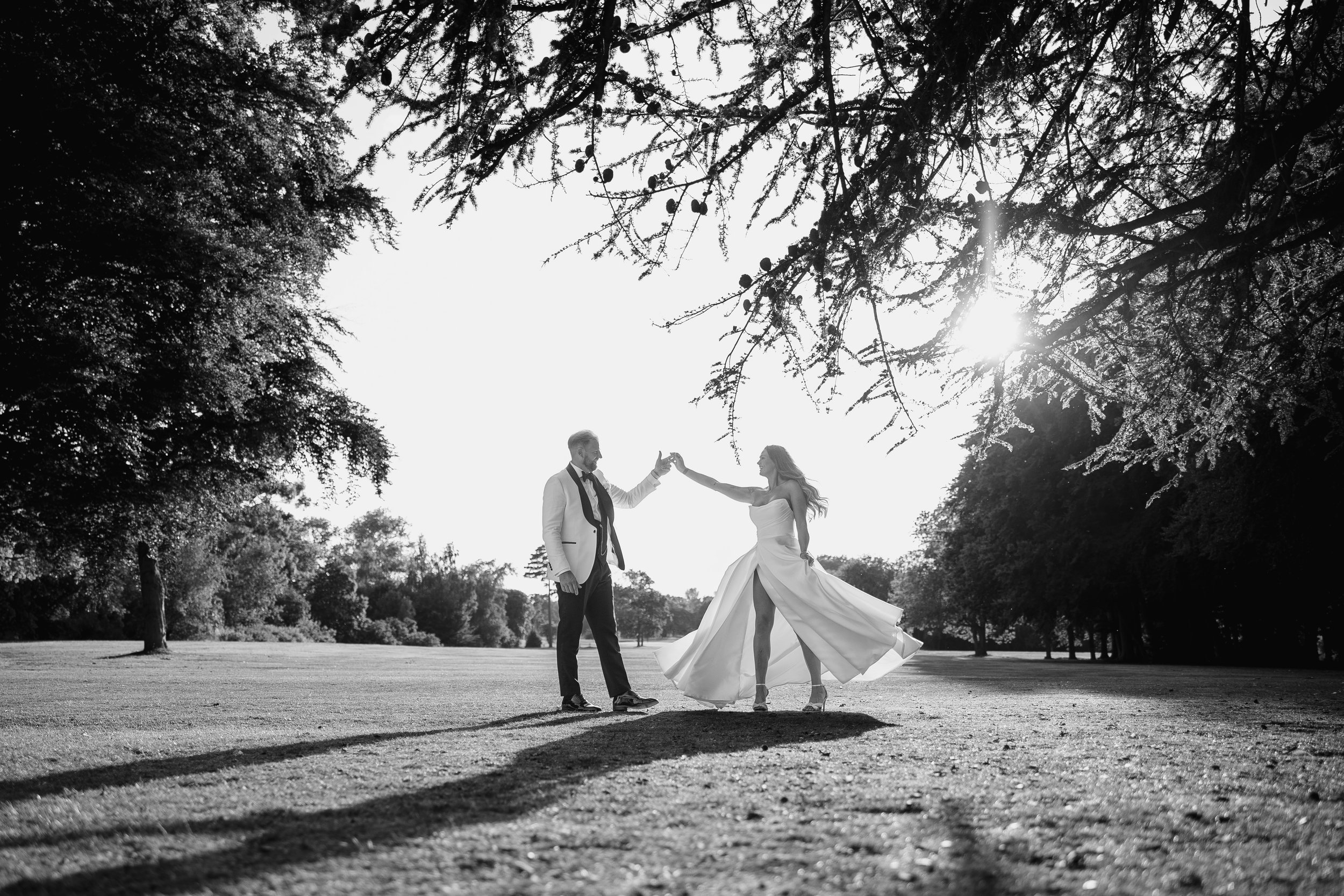 A black and white photo of a bride and groom holding hands and dancing outdoors under trees, with sunlight shining through the branches.