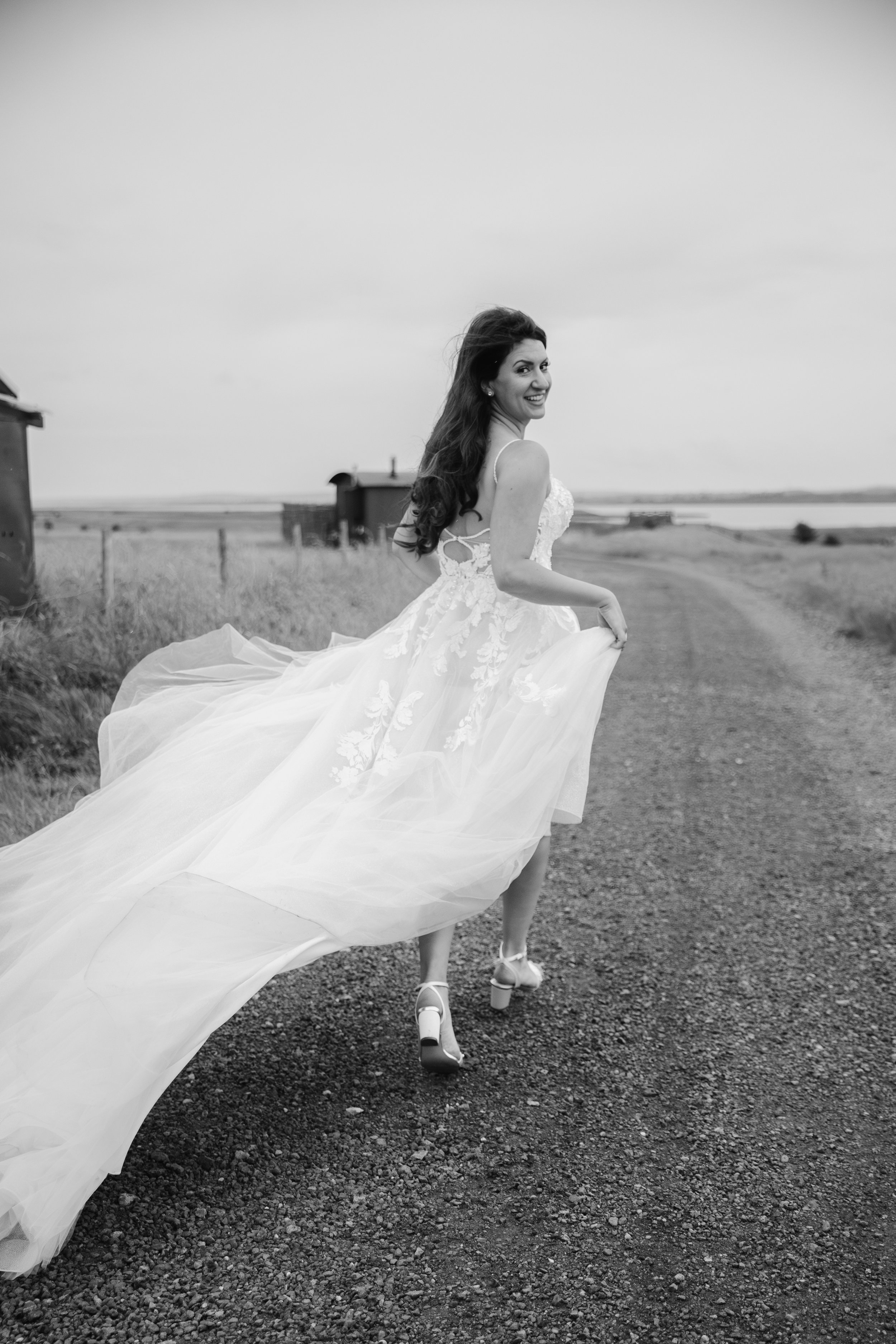 A woman in a wedding dress with a long train running on a gravel path in an outdoor landscape, smiling and looking over her shoulder.
