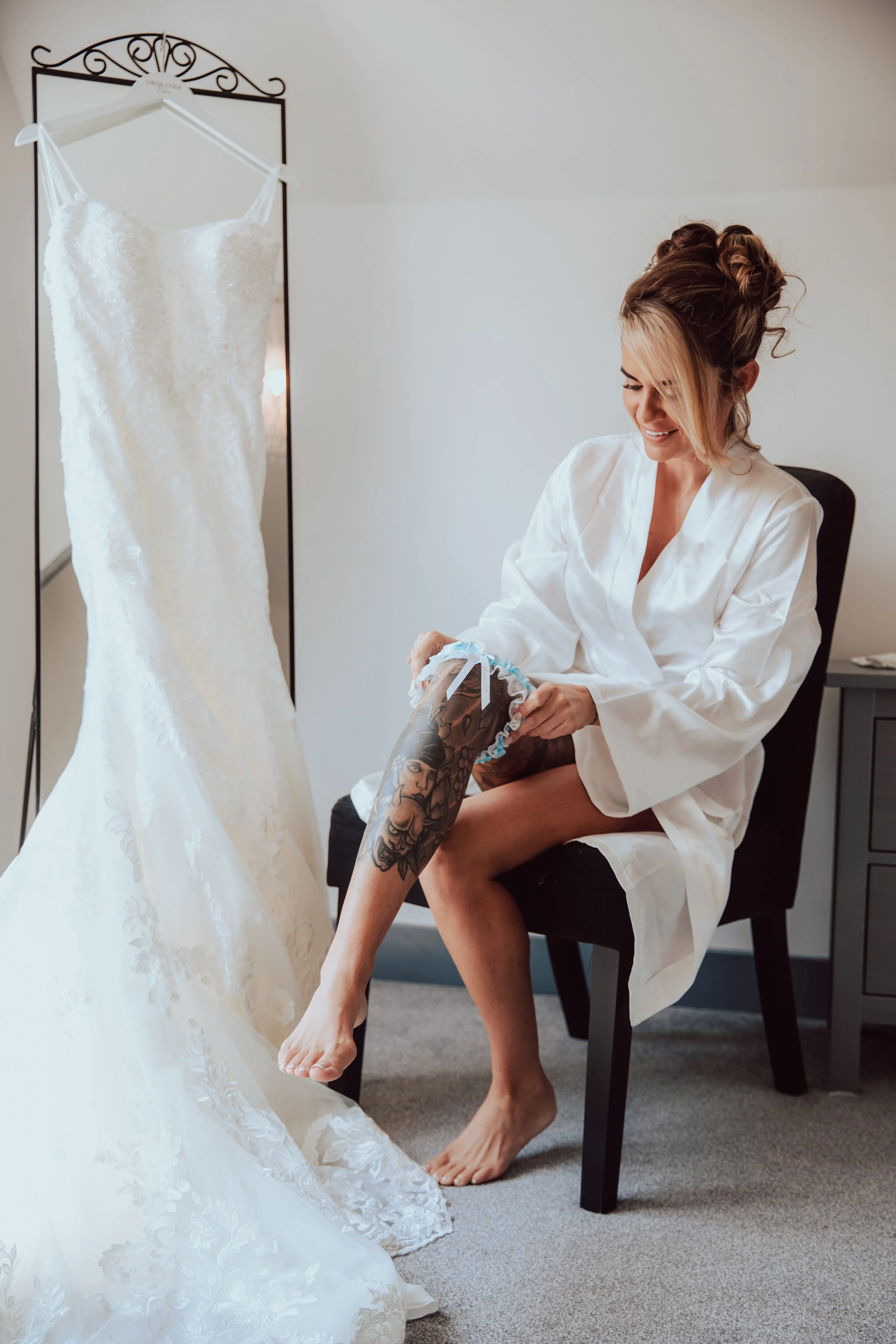 A woman with styled hair and tattoos putting on a shoe, sitting on a chair, with a wedding dress hanging on a mirror in the background.