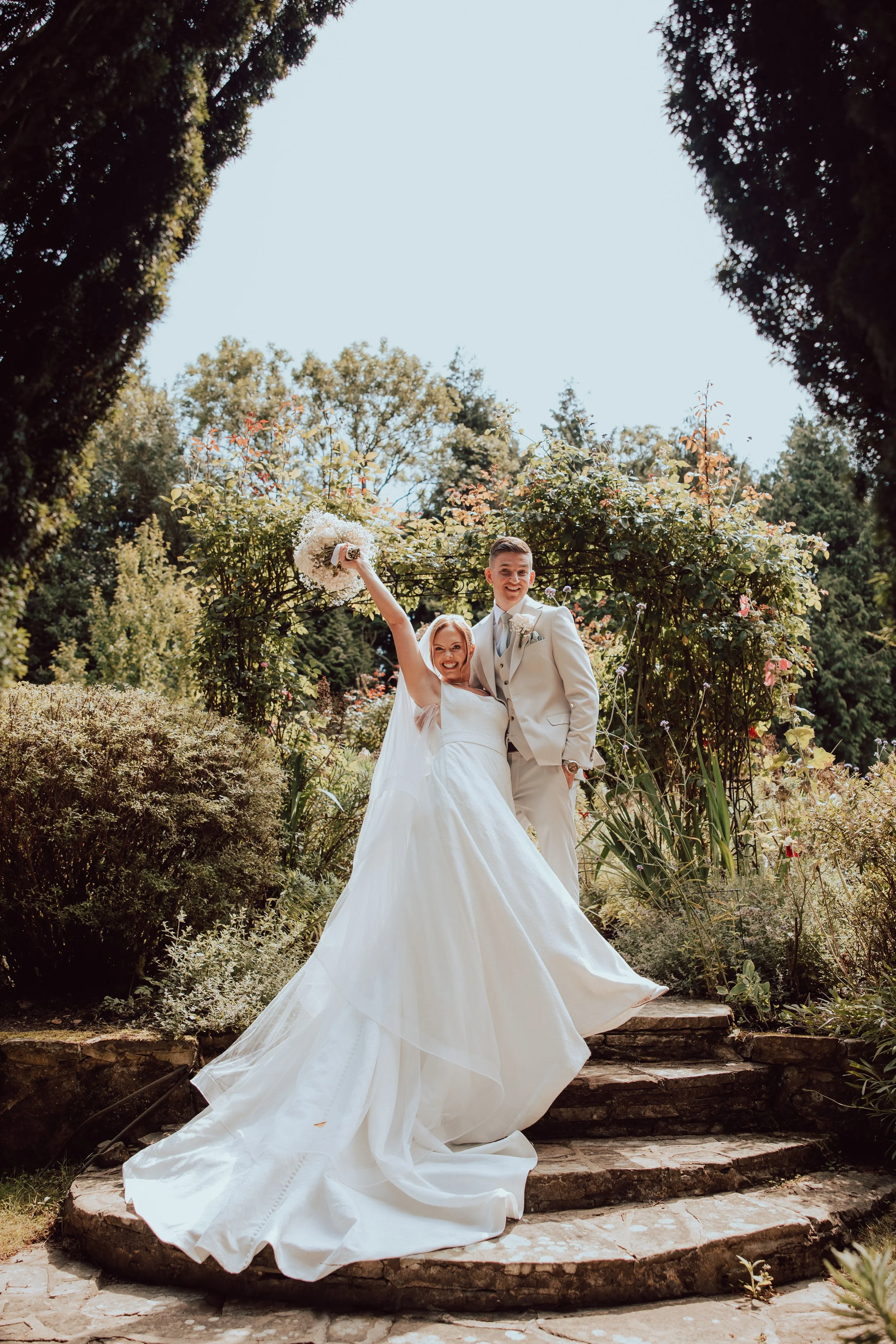 A bride and groom standing on stone steps in a garden with lush foliage and an arch of flowers behind them. The bride is holding a bouquet and smiling, while the groom is smiling and dressed in a white suit.