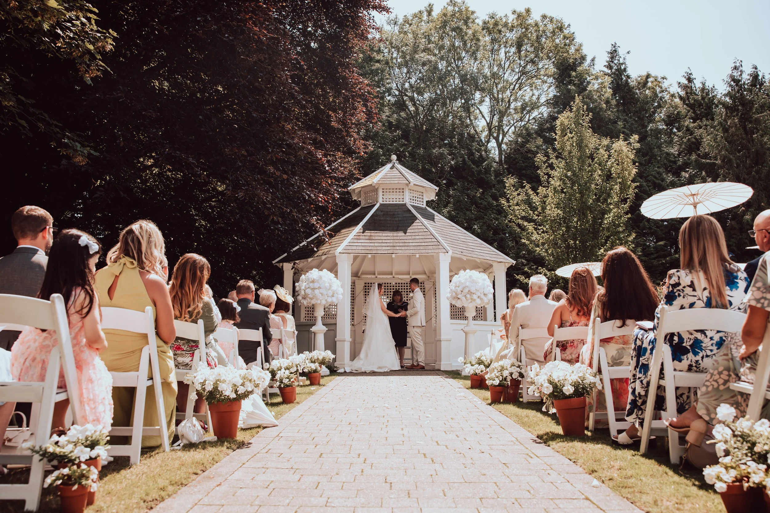 A wedding ceremony taking place outdoors under a gazebo with a bride and groom standing together, surrounded by seated guests and decorative white flowers along the aisle, with green trees and a clear blue sky in the background.