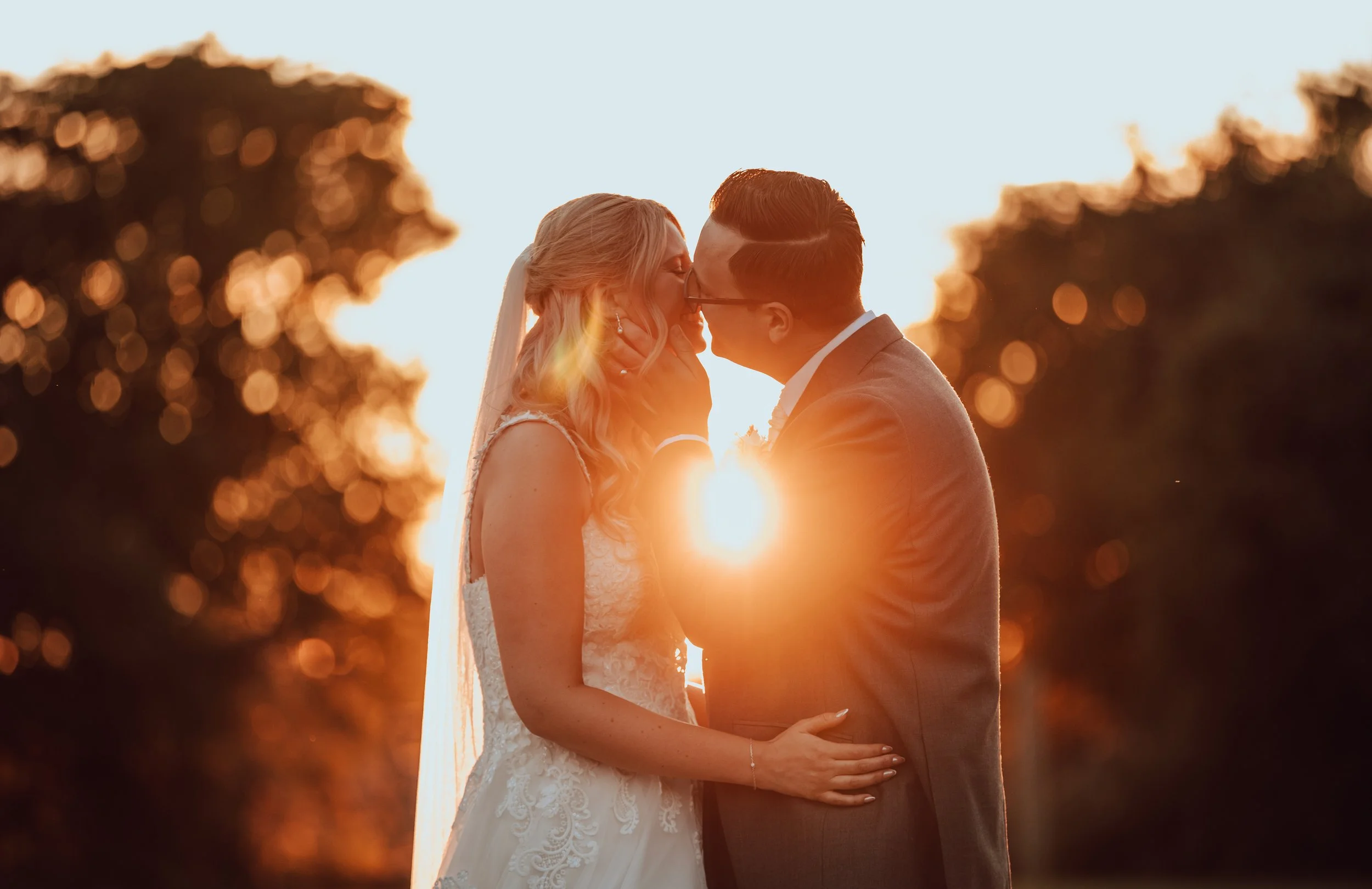 A bride and groom sharing a kiss at sunset, with the sun setting behind them and trees in the background.