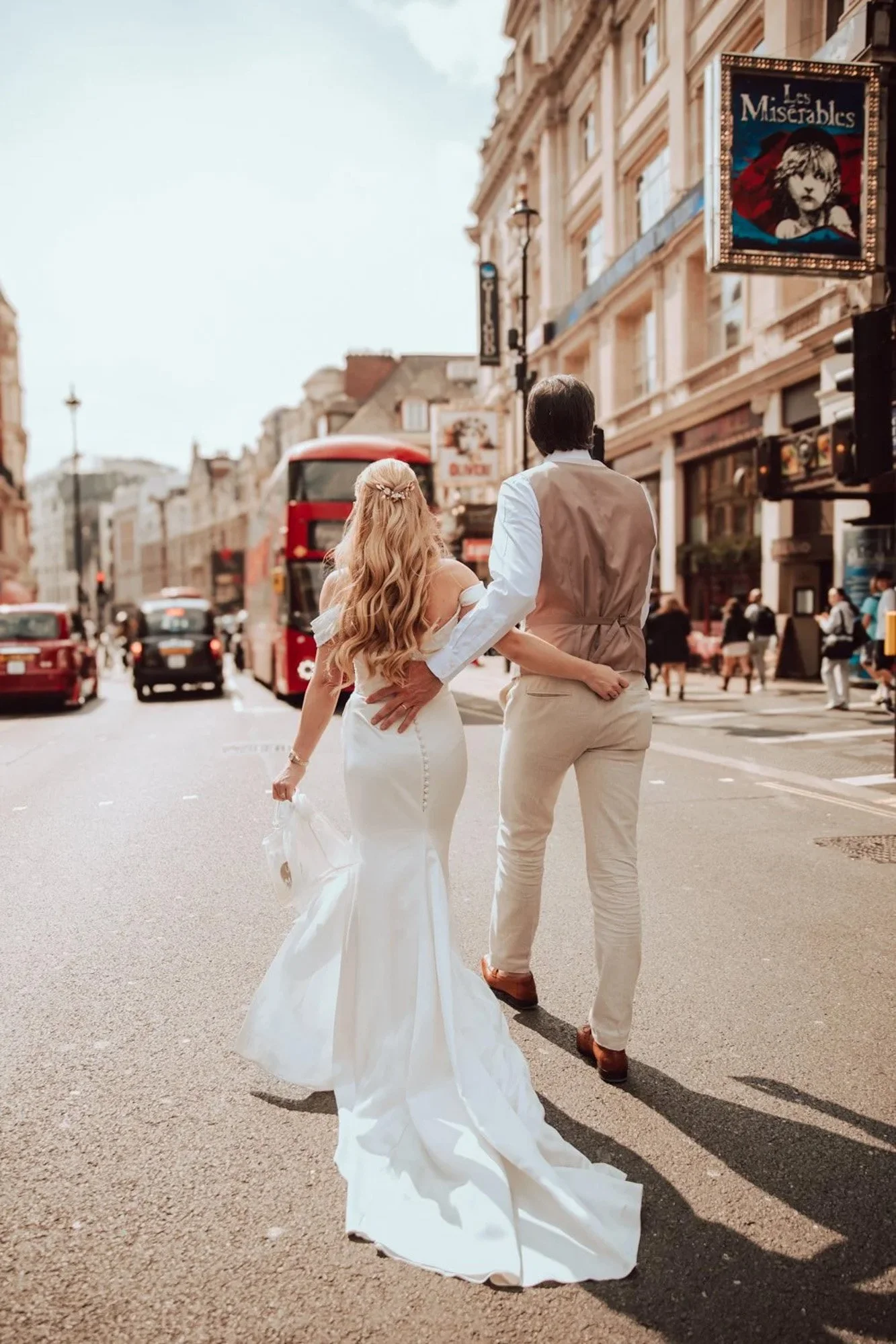 A bride and groom walking hand in hand on a city street, with the bride in a white wedding dress and the groom in beige trousers and a vest, surrounded by cars and pedestrians, with buildings and a billboard for Les Misérables in the background.