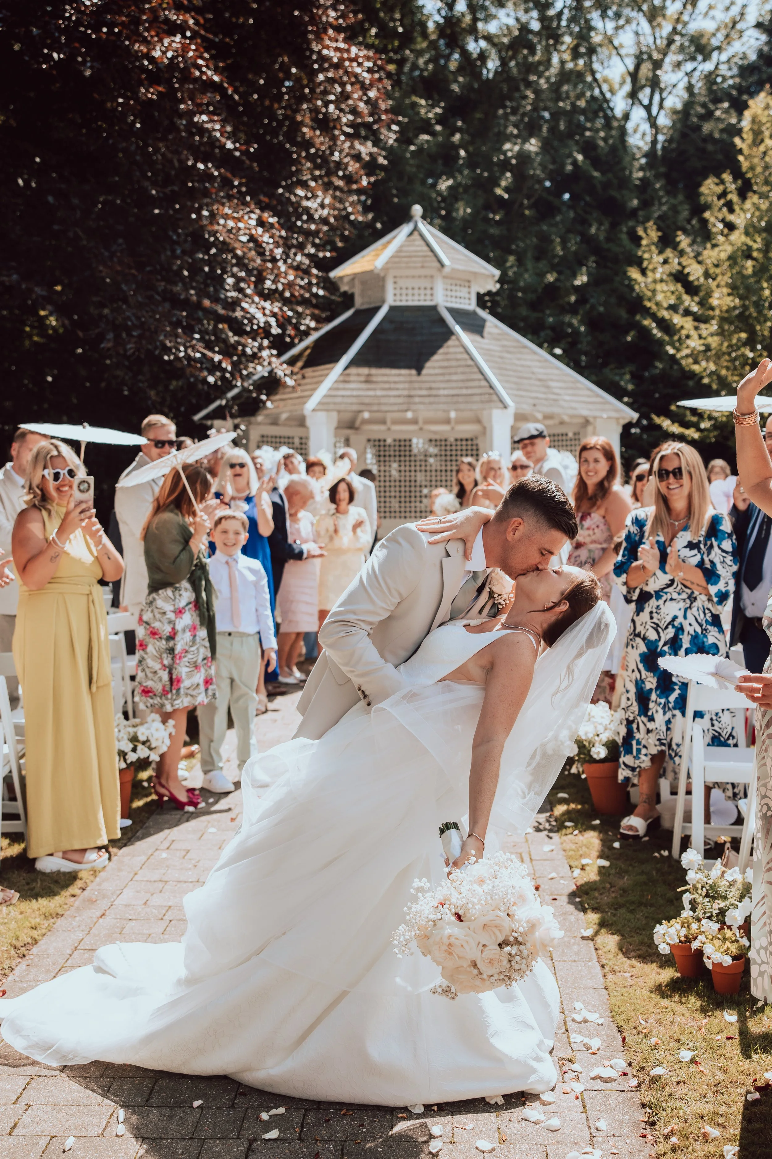 A wedding scene outdoors with the bride and groom sharing a kiss, surrounded by guests. The bride is in a white gown, holding a bouquet, and the groom in a tuxedo. Guests are smiling, clapping, and taking photos, with some holding umbrellas. There is a small pavilion in the background and flower arrangements along the aisle.
