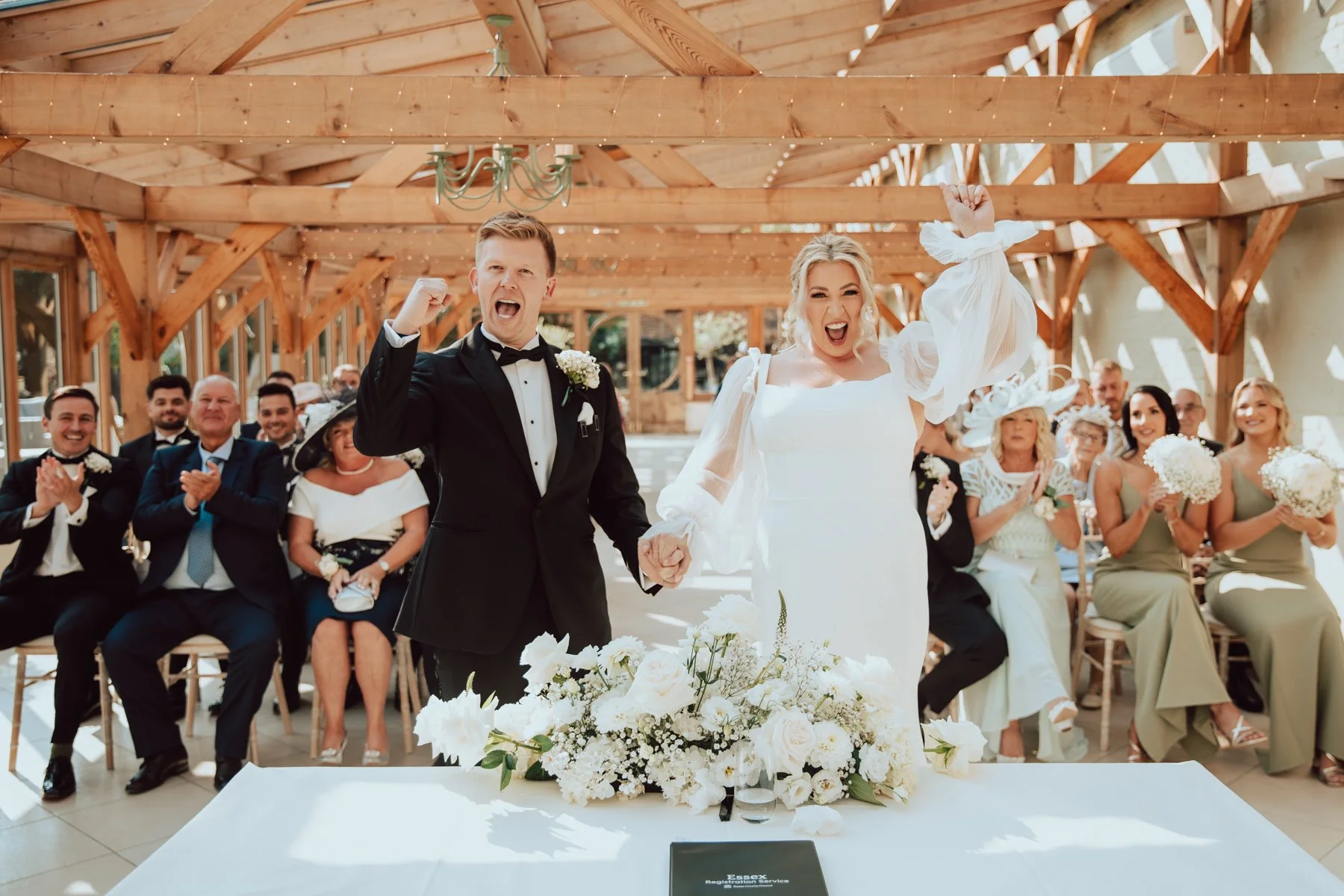 A newly married couple dressed in wedding attire celebrating during their wedding ceremony. The groom is in a black tuxedo, and the bride is in a white wedding dress holding hands and smiling. Guests are seated behind them, clapping and smiling. The setting is indoors with a wooden ceiling and natural light.