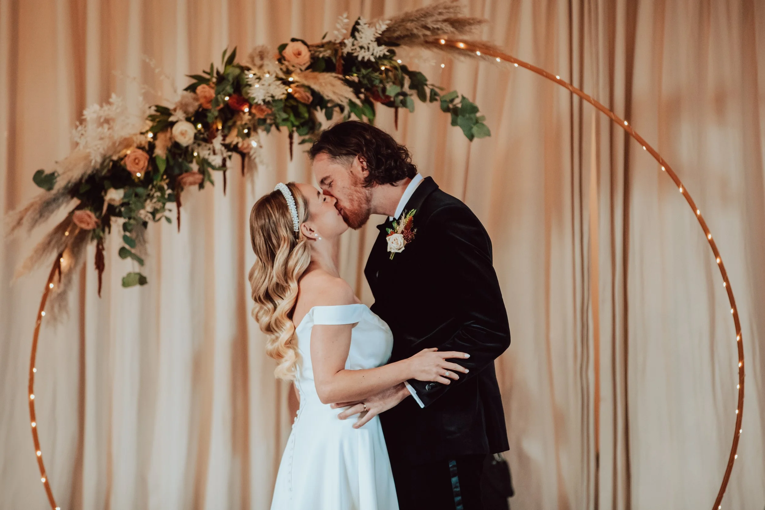 A bride and groom kiss during their wedding ceremony, with a floral arch decorated with roses, greenery, and pampas grass in the background.