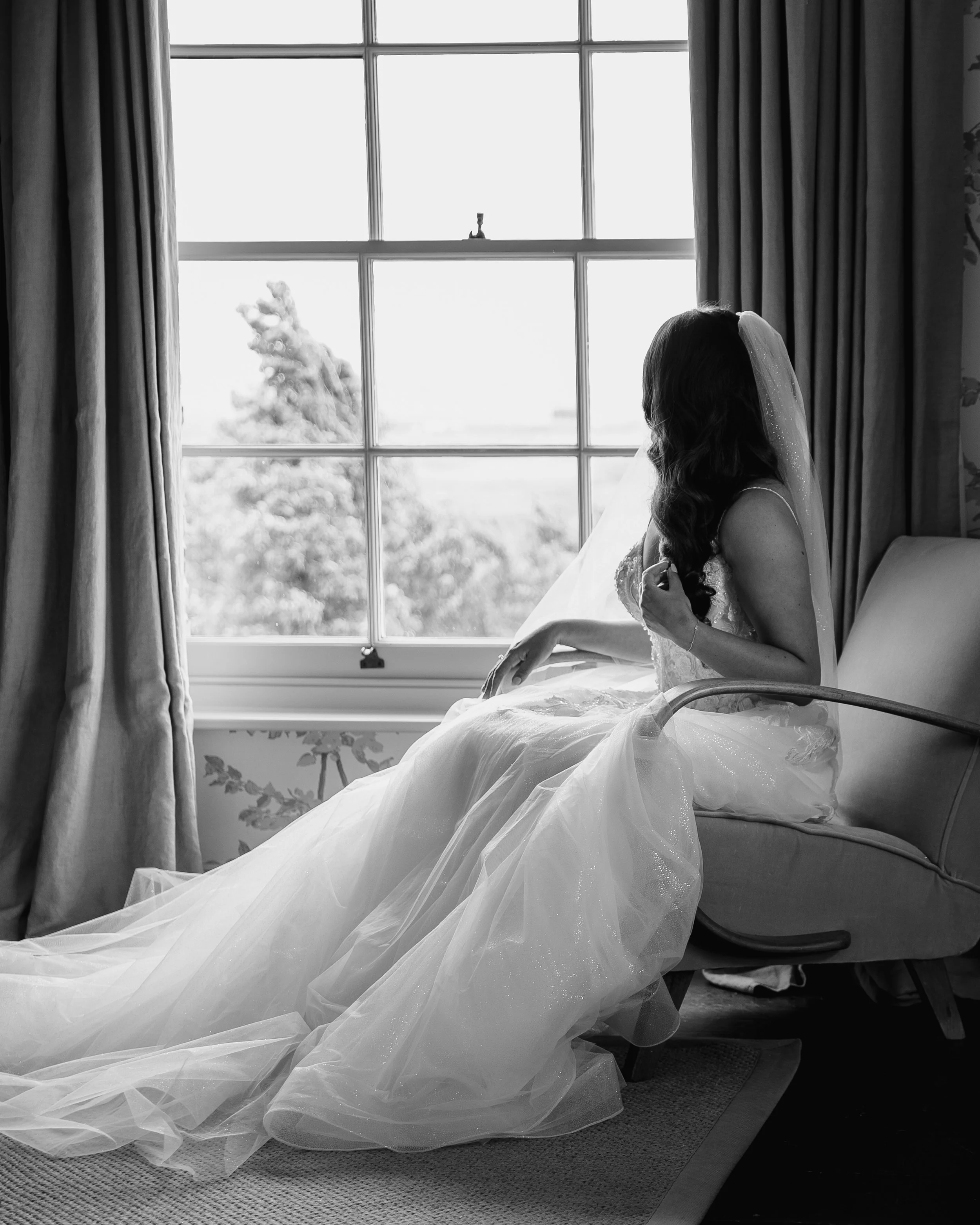 A woman in a wedding dress sitting beside a large window with curtains, looking out, with her wedding veil and dress flowing around her.