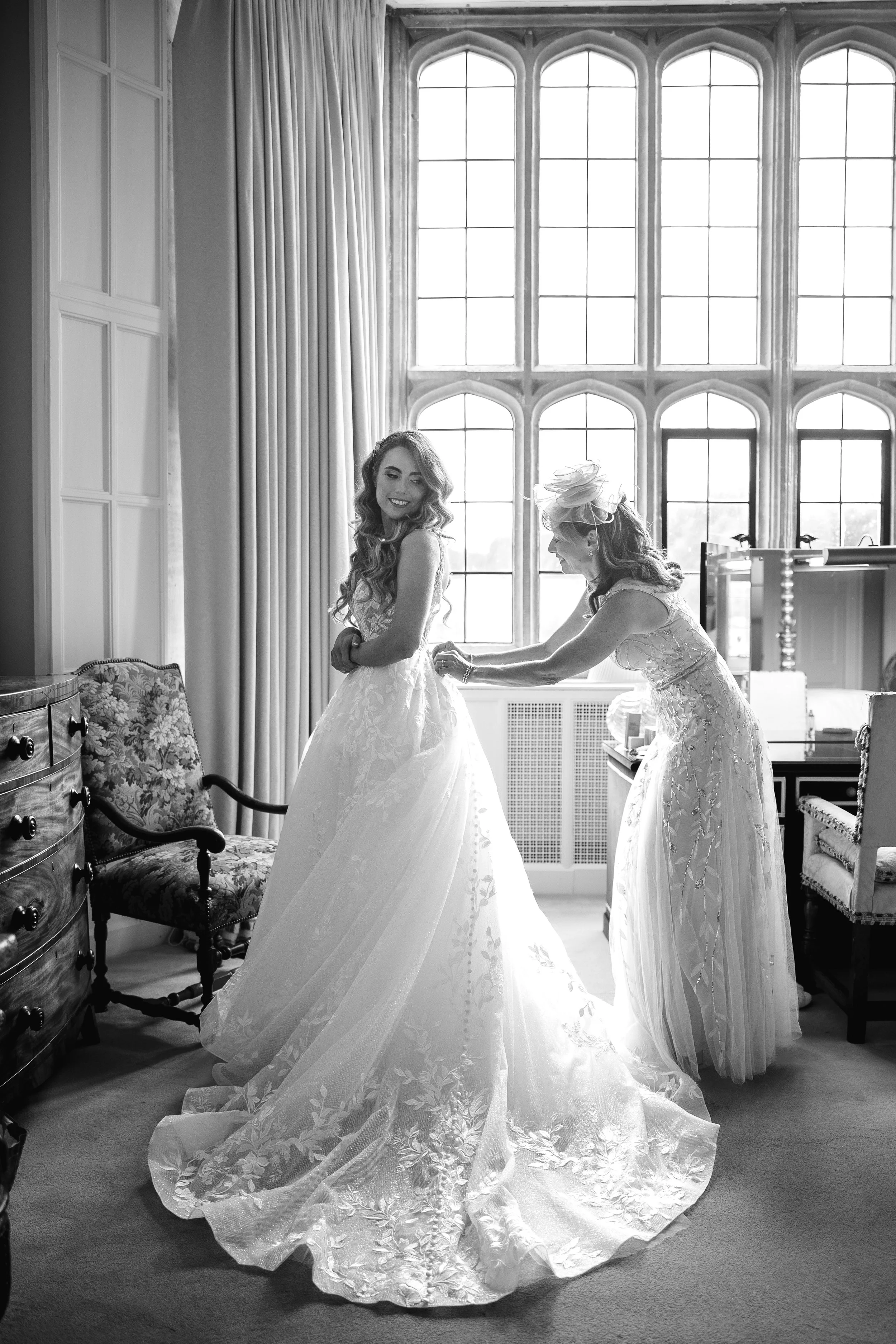 A woman in a wedding dress smiling as another woman, likely a bride's attendant, helps adjust her gown in a room with large windows and elegant furniture.
