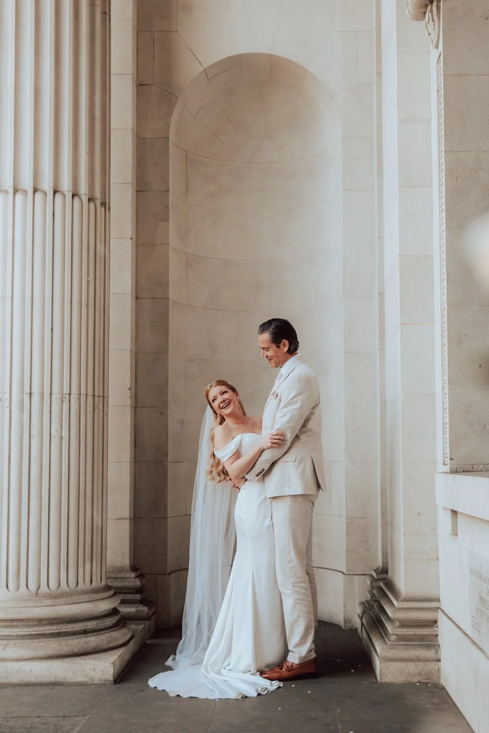 A bride and groom in wedding attire sharing a joyful moment inside a classical building with large columns and stone walls.