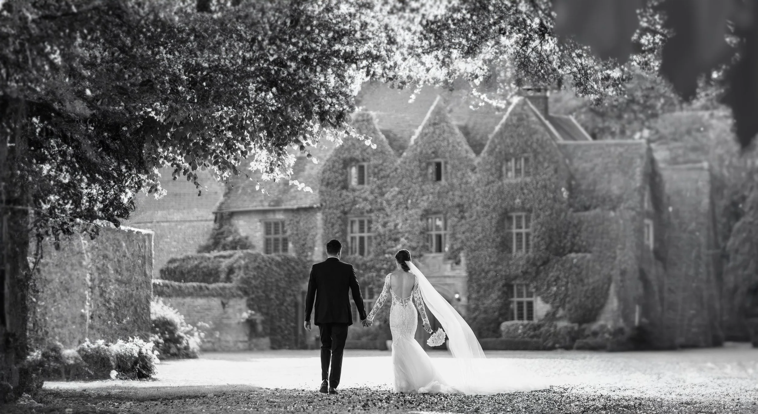A bride and groom walking hand in hand outdoors near a large house, with trees and greenery around, captured in black and white.