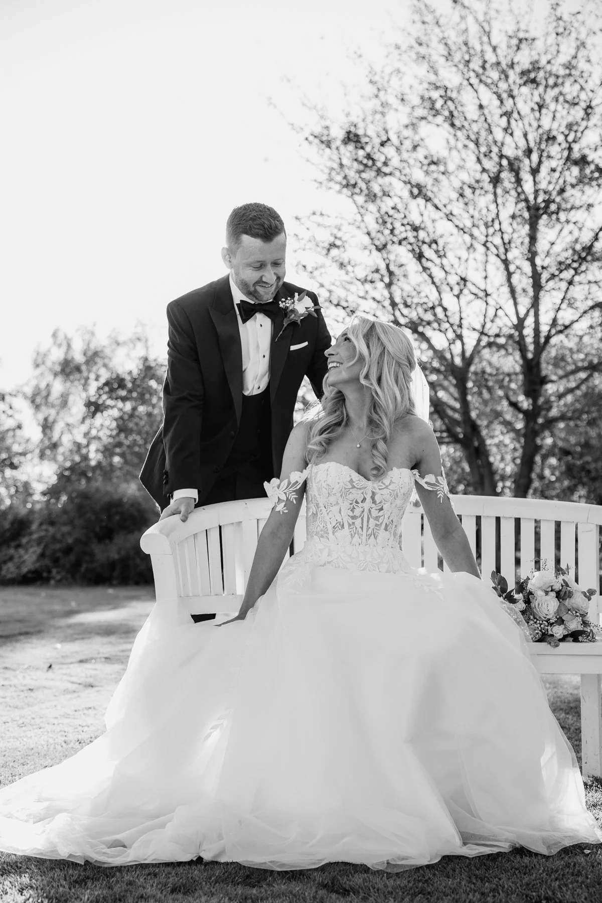 A black and white photo of a bride in a wedding dress sitting on a bench with a bouquet, and a groom in a tuxedo standing beside her outdoors, smiling.