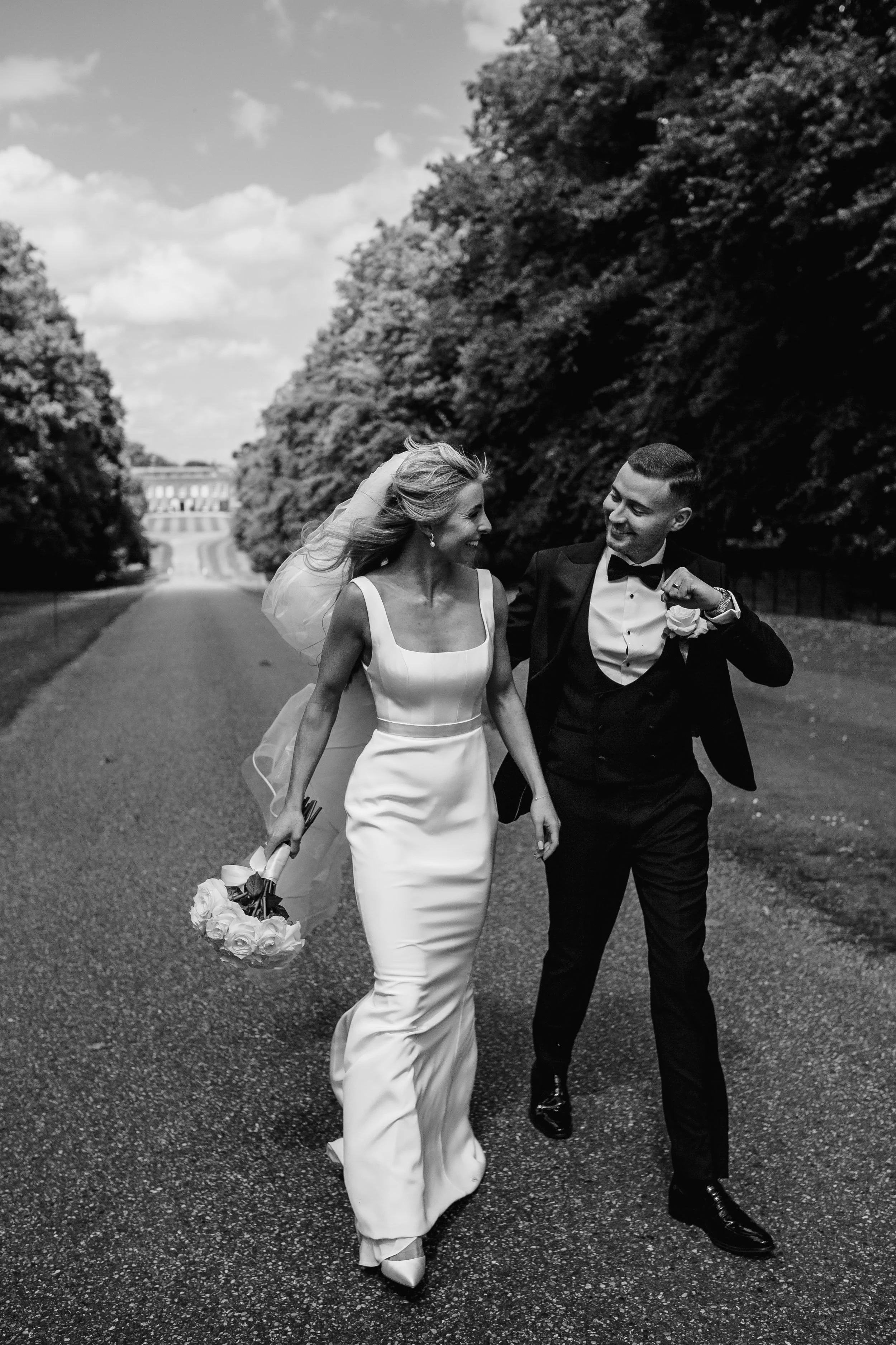 A black and white photo of a bride and groom walking hand in hand outdoors. The bride wears a white wedding dress and holds a bouquet, while the groom is in a black tuxedo with a bow tie and a boutonniere. They are smiling at each other.